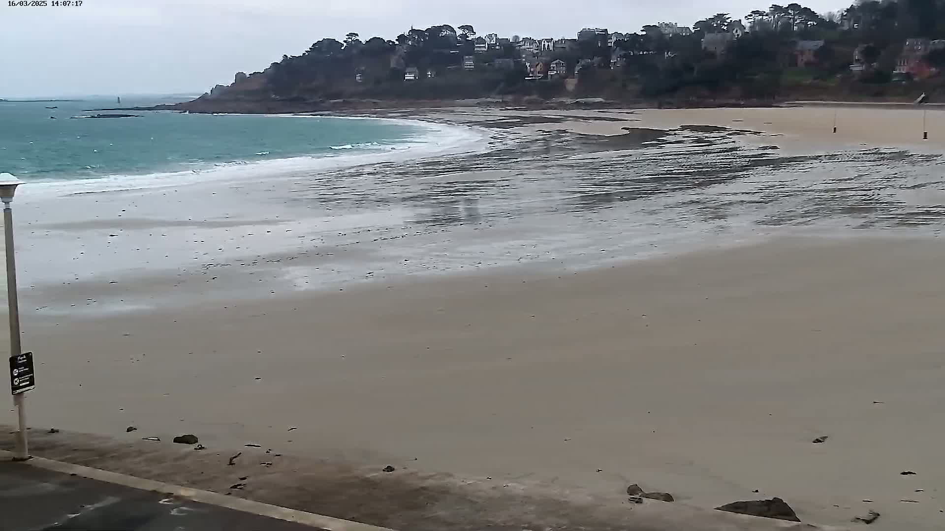 A wide sandy beach with gentle waves under an overcast sky, backed by a line of houses nestled amongst trees on a cliff.
