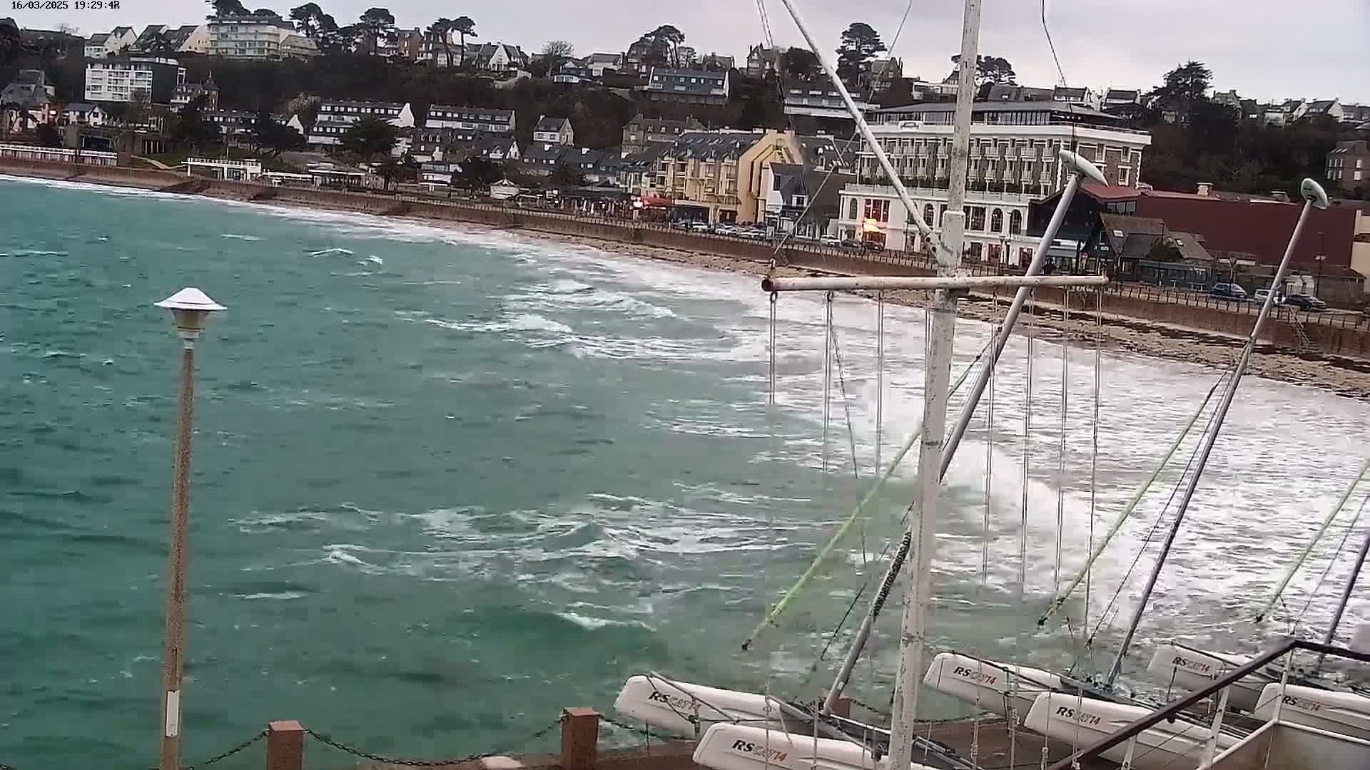 Several small sailboats are partially submerged in choppy, green-tinged water near a beach and waterfront buildings under an overcast sky.