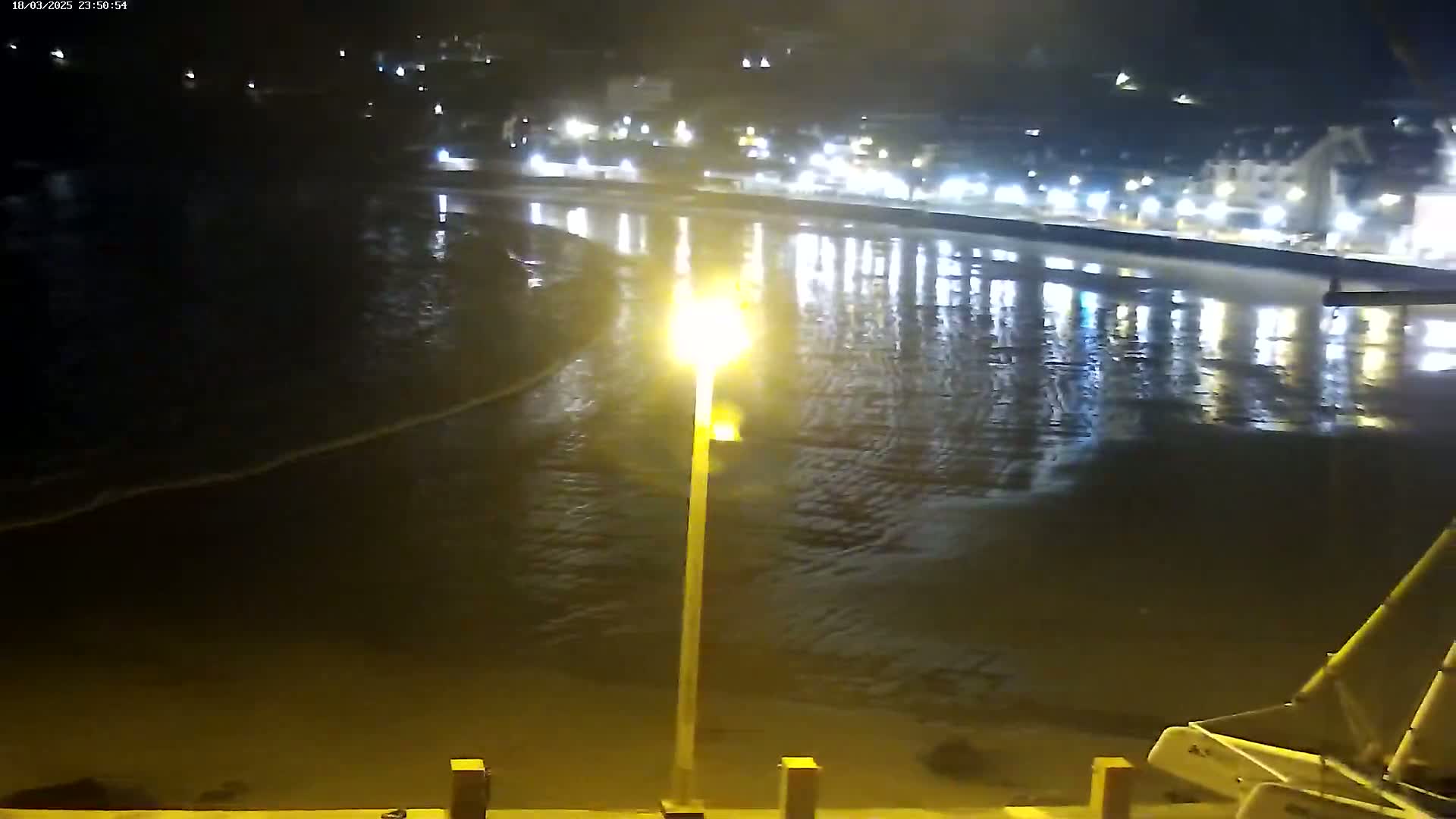 A nighttime view of a calm body of water reflecting the lights of a town on the shore, with a sailboat visible in the lower right corner.