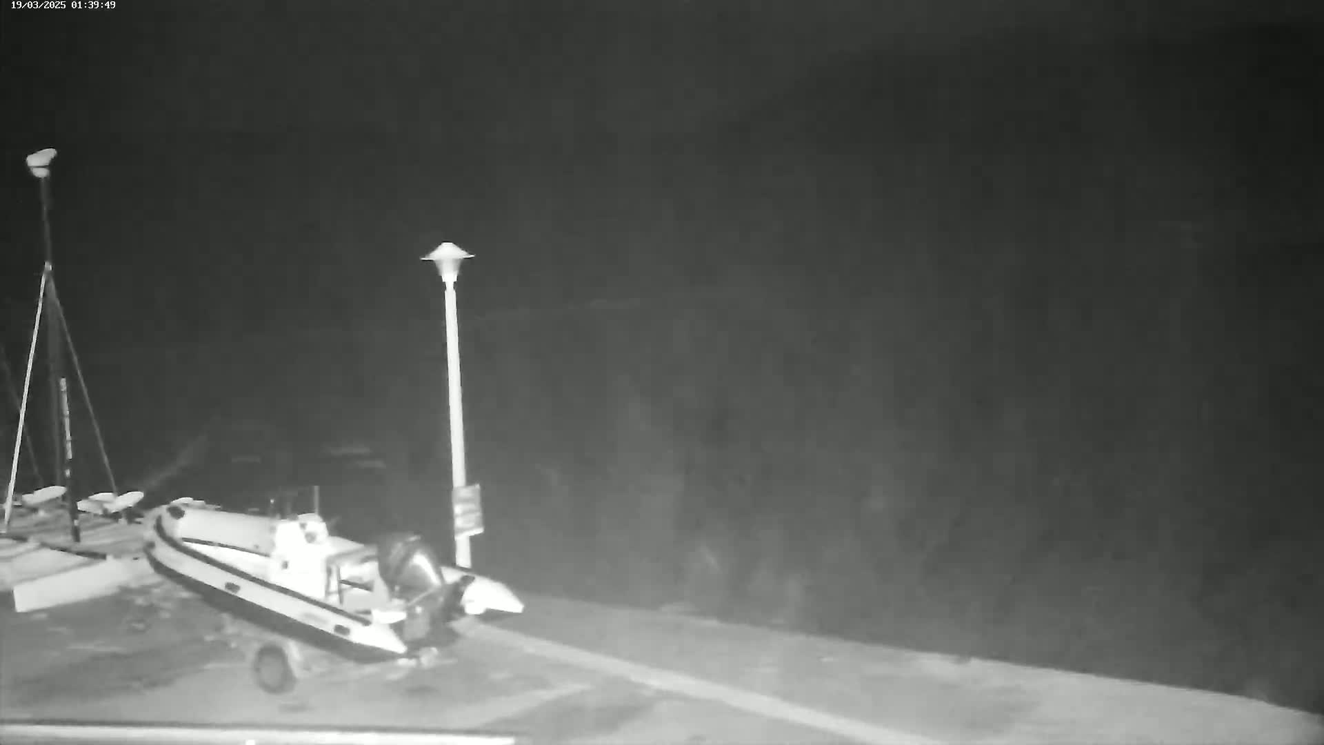 A rigid-hulled inflatable boat is parked on a dark, nighttime dock next to a tall lamppost and another, smaller structure.