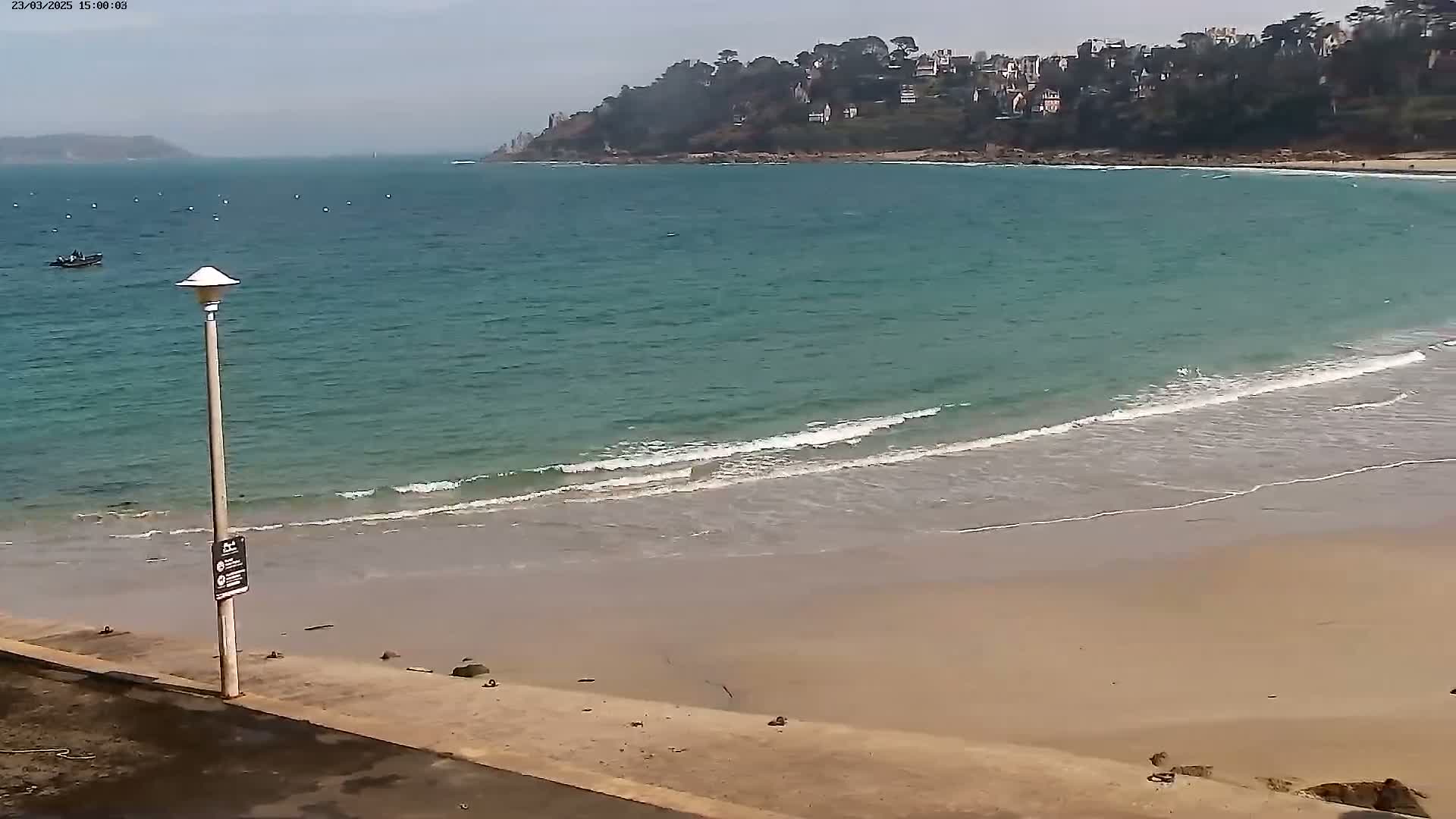 A sandy beach with gentle waves laps the shore under an overcast sky, with a small town nestled on a hill in the distance.