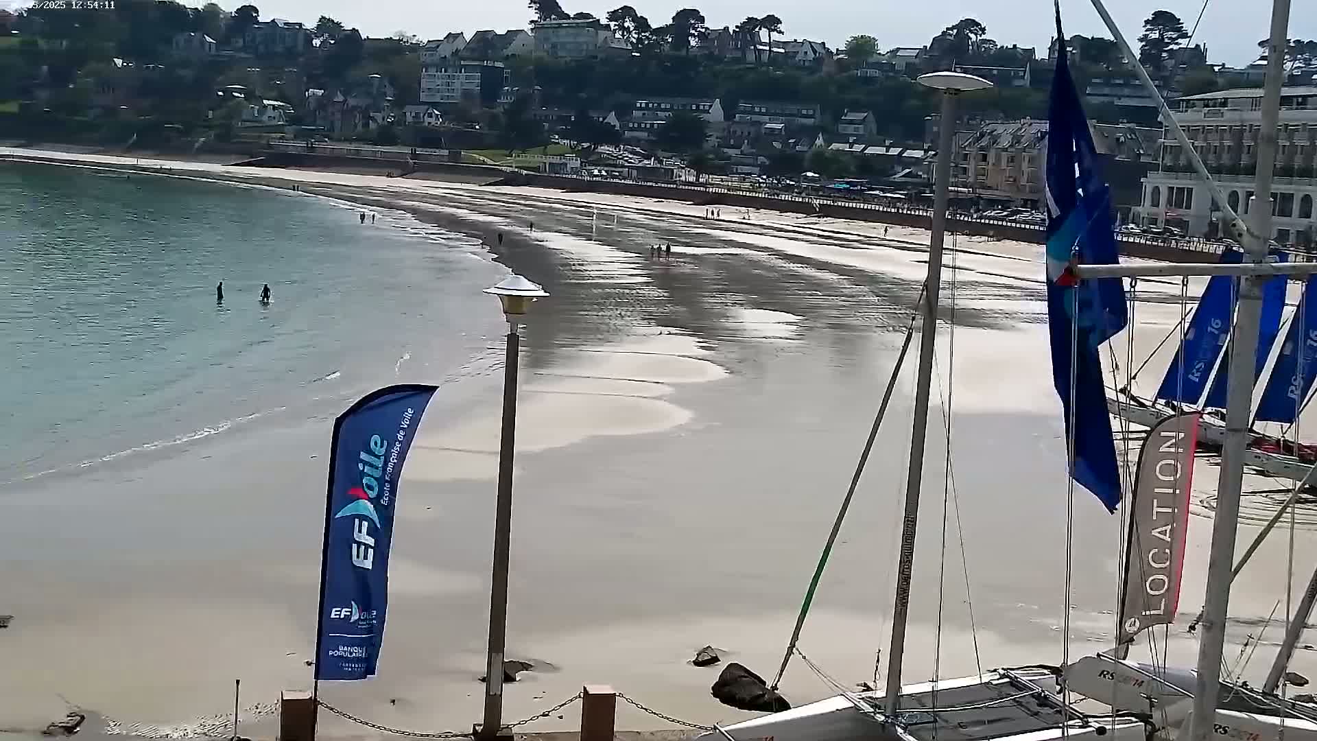A mostly sunny day shows a sandy beach with a few people walking and wading in the shallow water, next to a calm bay with sailboats moored nearby and a town rising up on the hill behind.