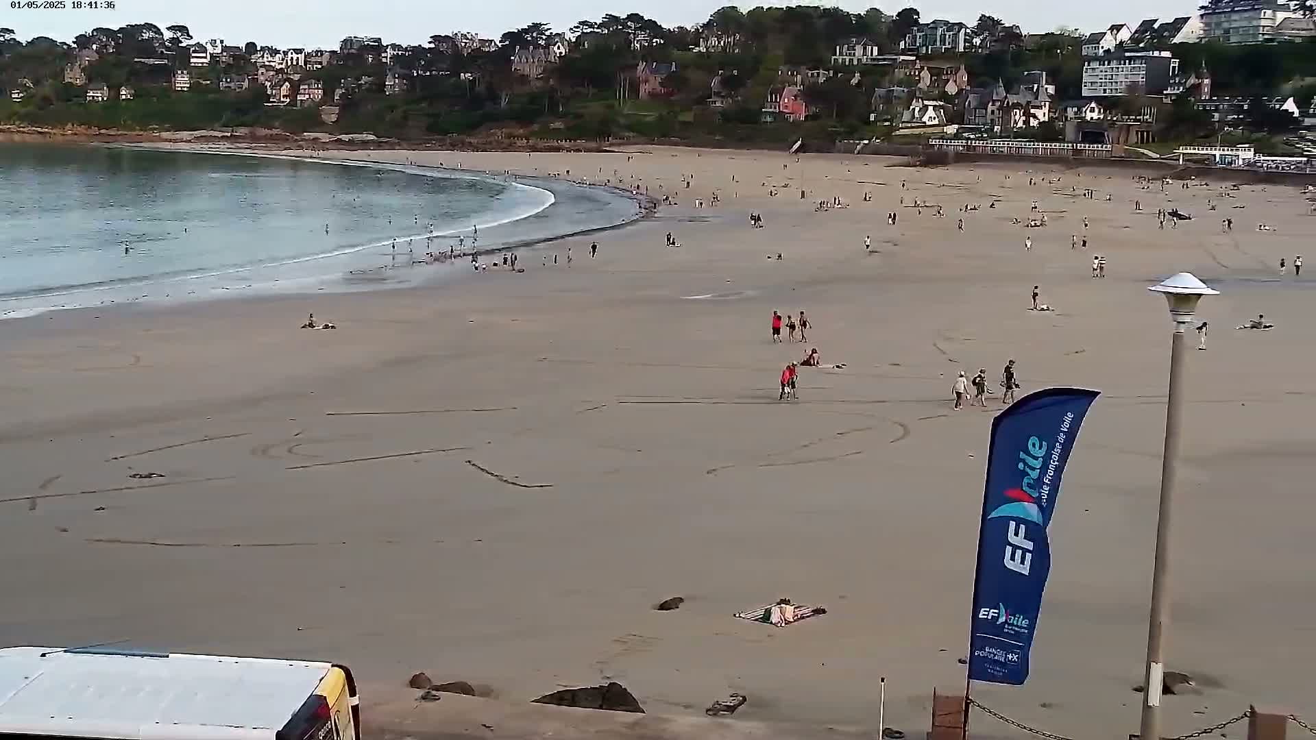 A sandy beach with many people scattered across it, gently sloping toward calm ocean waves, is situated below a line of houses and trees on a partly cloudy day.