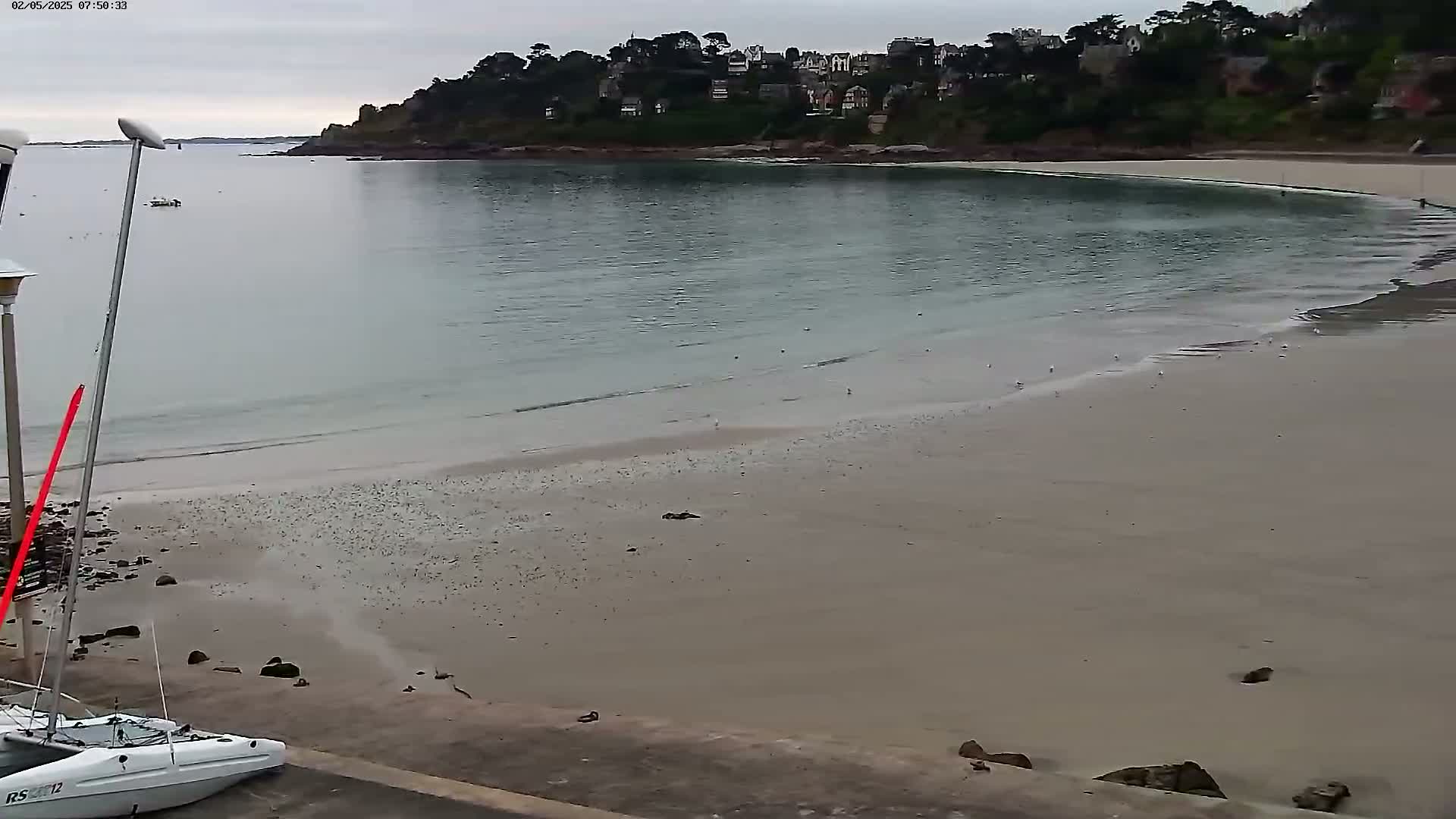 A sailboat is moored near a sandy beach, overlooking a calm bay with a town nestled on a hillside under an overcast sky.