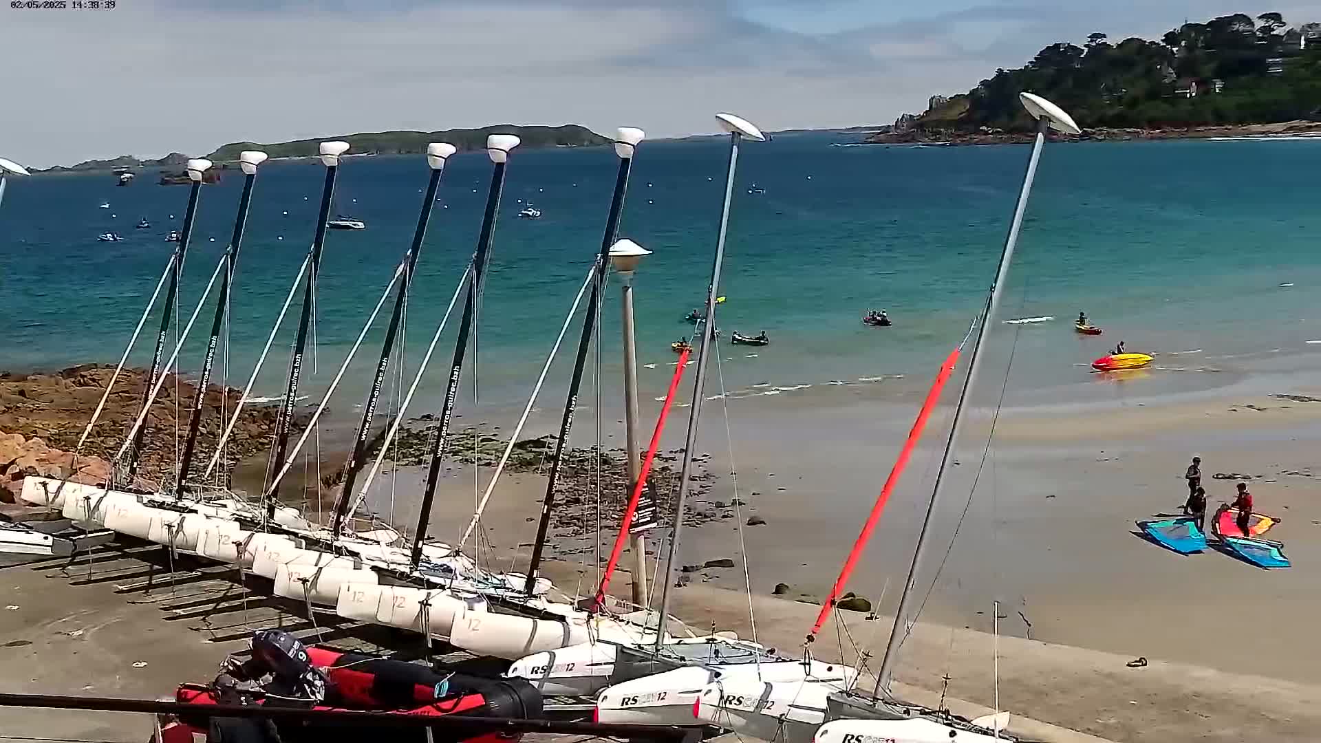 Several small sailboats are beached on a sandy shore next to a calm, turquoise ocean under a partly cloudy sky; several people are visible on the beach and in the water.