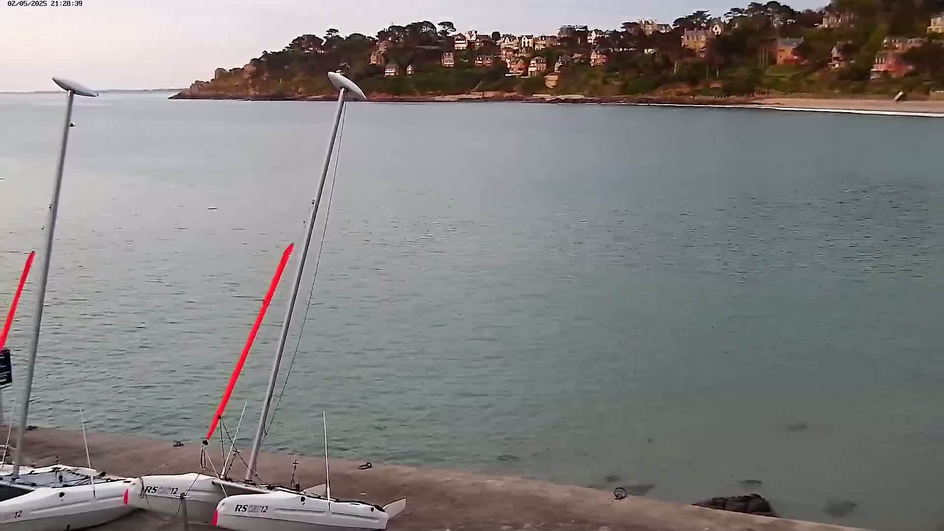 Two sailboats are docked on a concrete shore next to calm, gray-green water, with a residential area visible across the bay under an overcast sky.