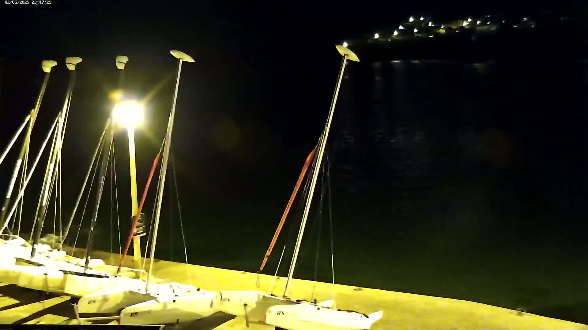 Several sailboats are docked at a waterfront at night under bright lights, with calm dark water visible beyond.