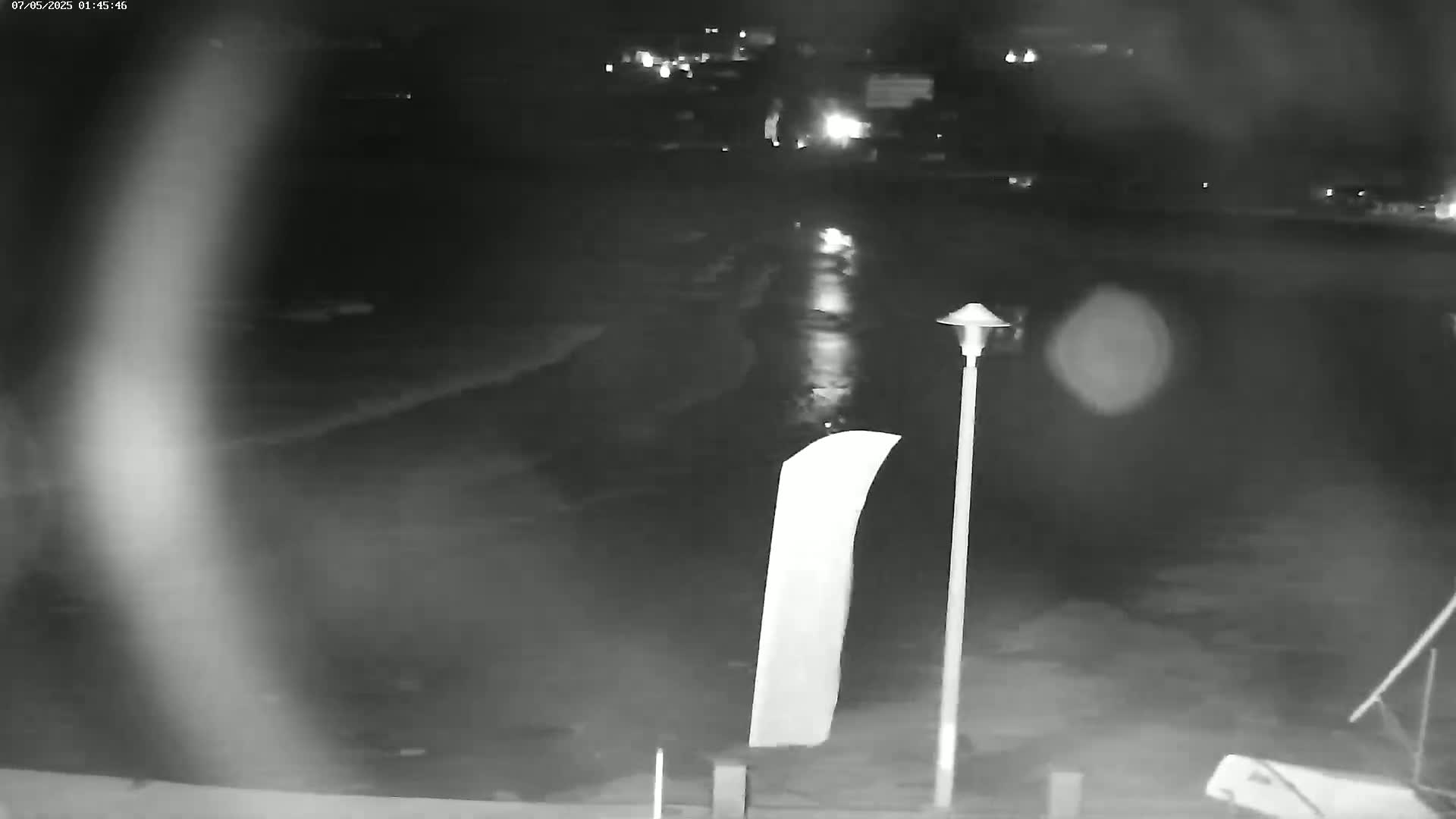A nighttime, low-light outdoor image shows a wet, dark paved area with a tall, slender lamppost and a large, light-colored sign.