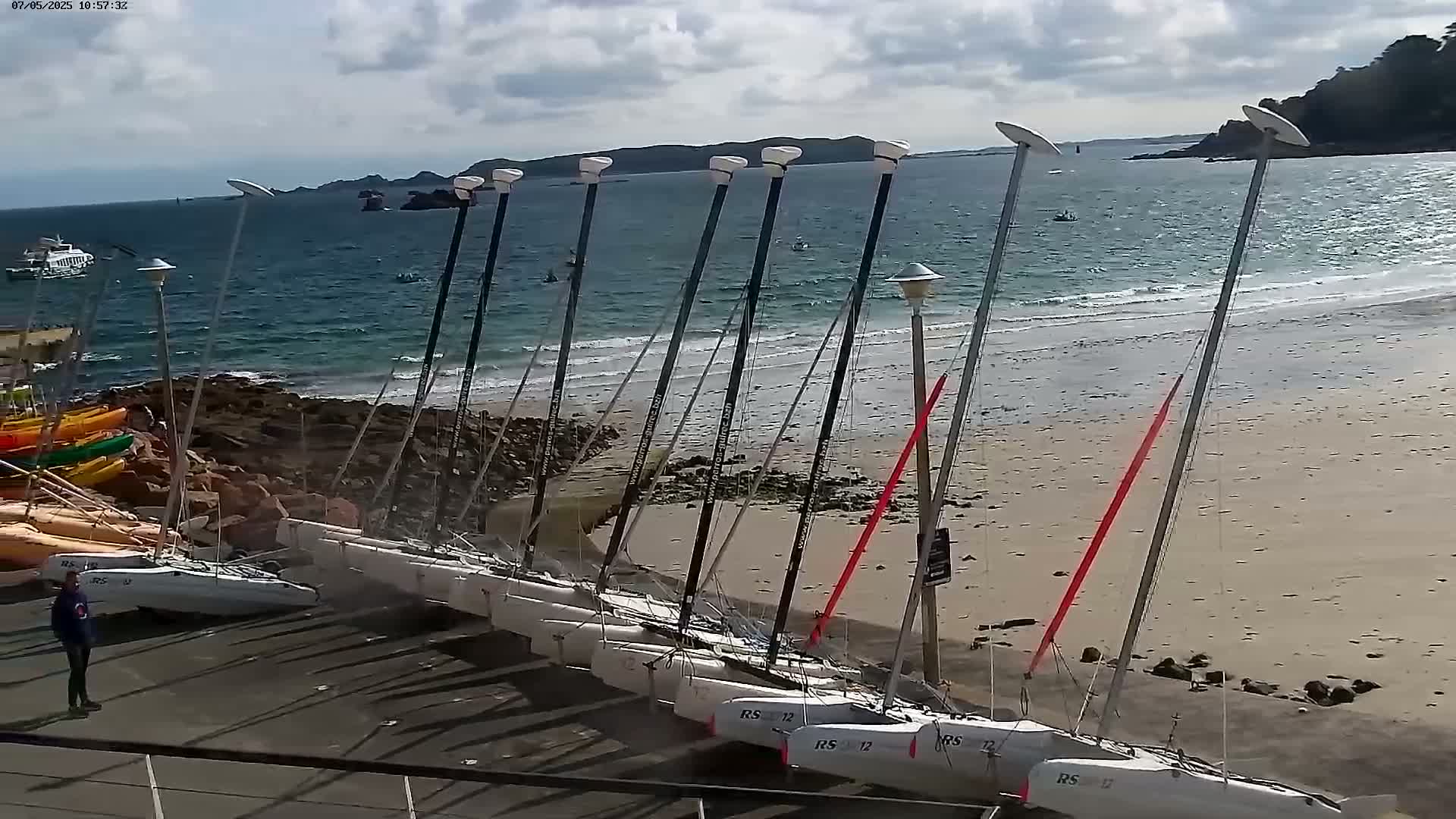 Numerous sailboats are beached on a sandy shore near rocky outcrops, under a partly cloudy sky, with calm water visible beyond.