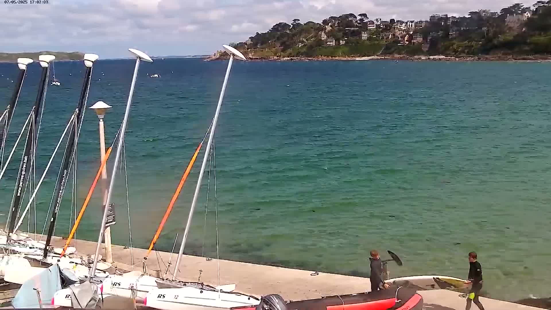 Several sailboats are moored at a quay next to teal-colored water, and two people stand near a surfboard on the shore under a partly cloudy sky.