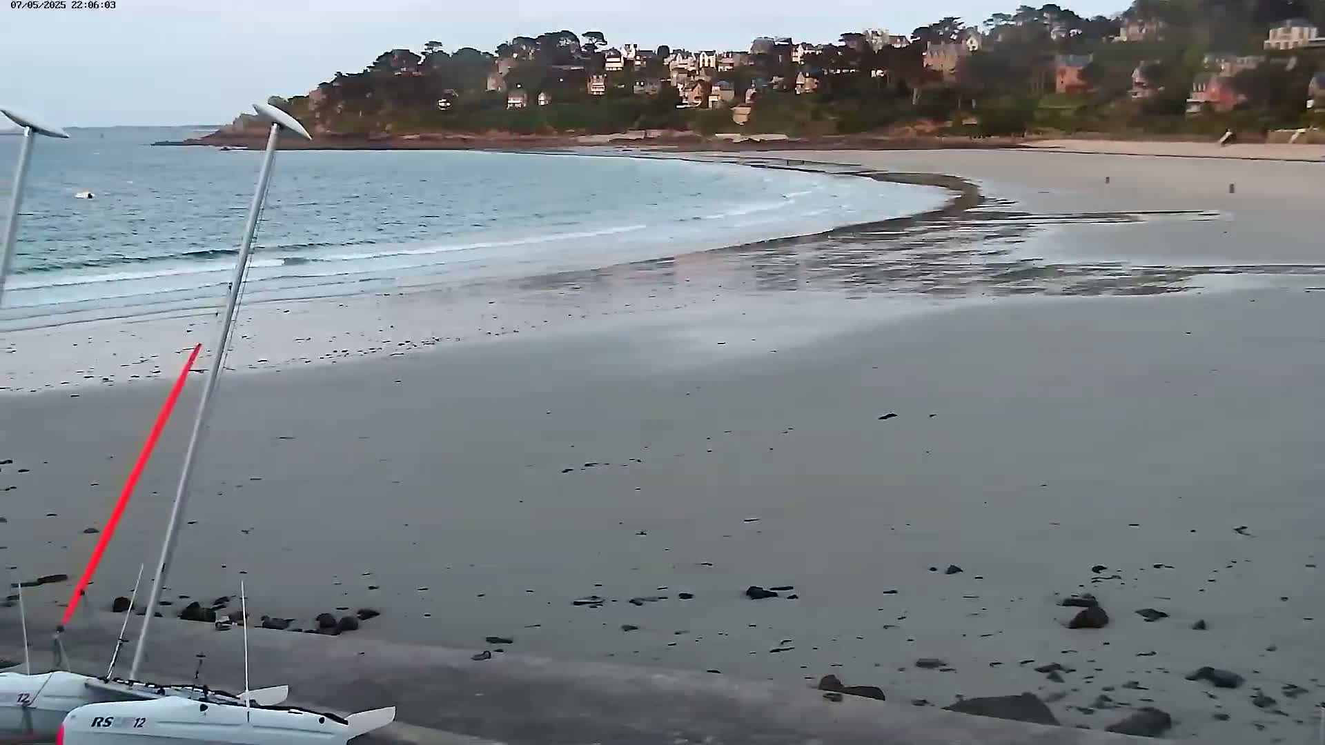 A sailboat is beached on a sandy shore next to calm, light-blue water, with a residential area visible on a hill in the background under an overcast sky.