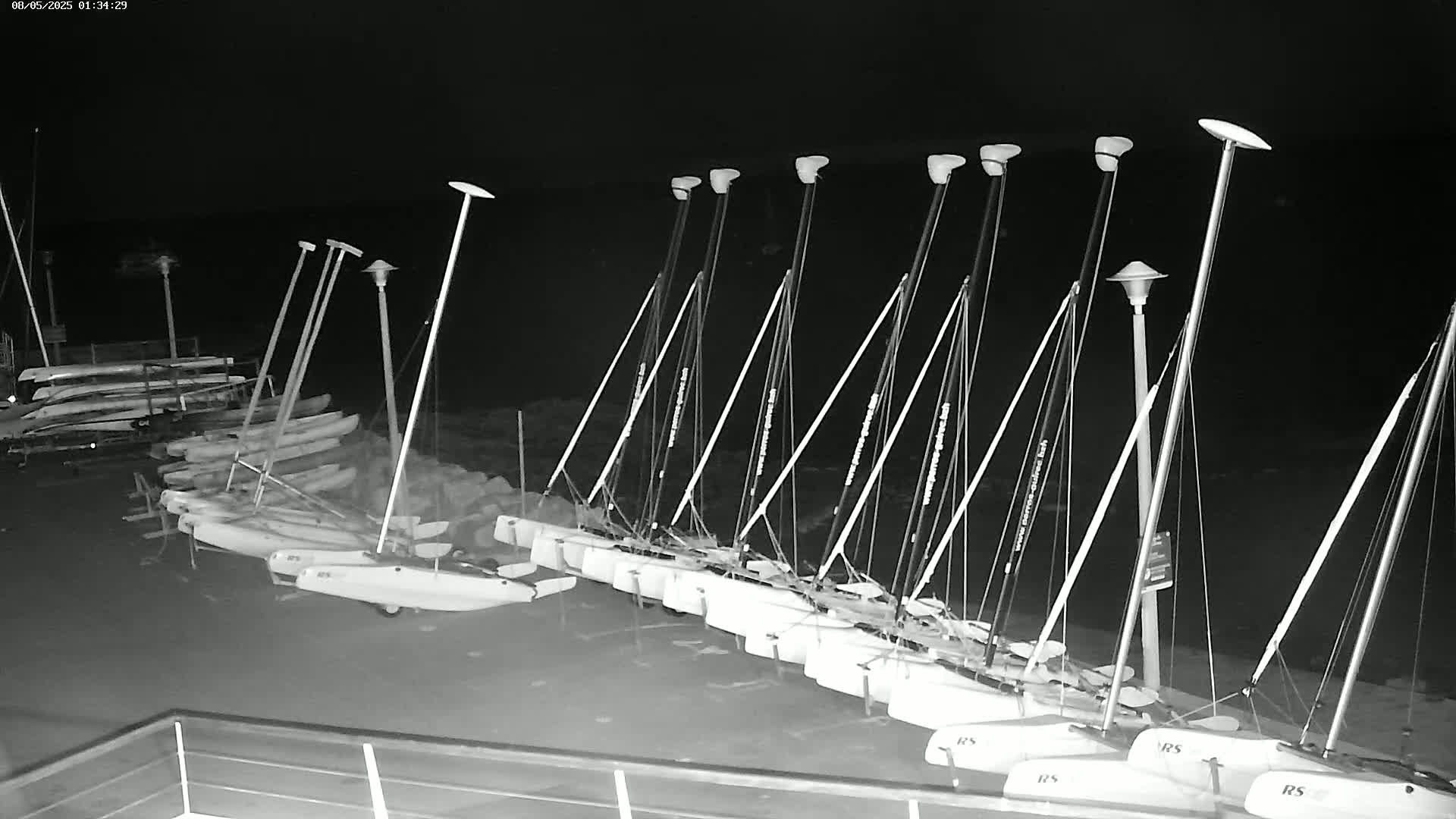 A row of small sailboats is beached at night under several tall lights.