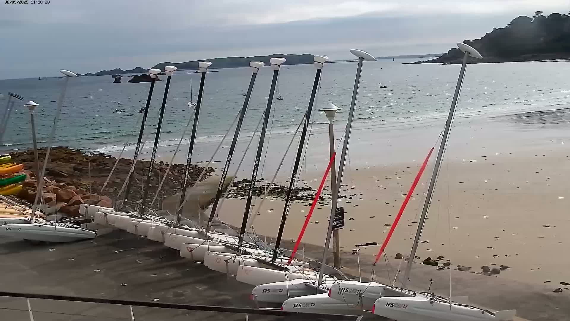 Several catamarans are beached on a sandy shore next to rocky outcrops, under a cloudy sky, with calm water visible in the background.