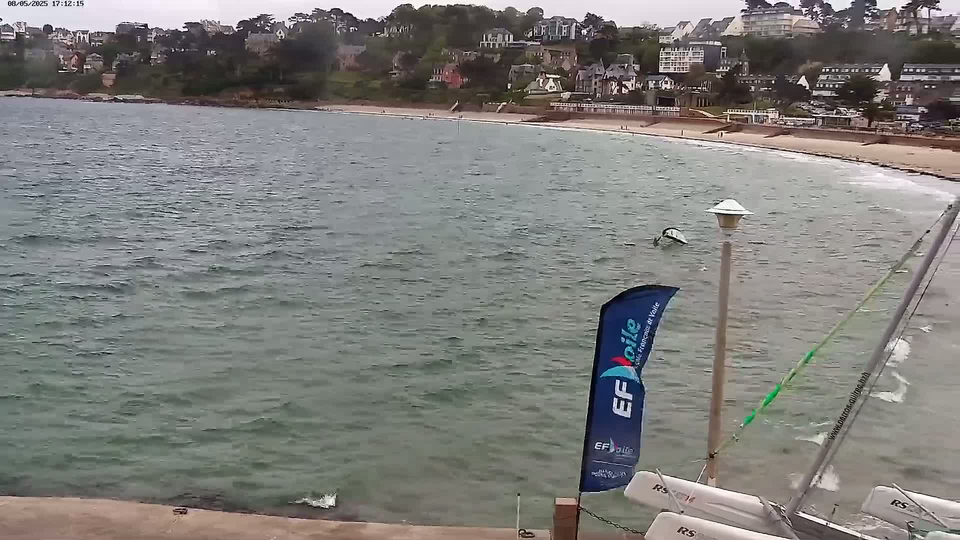 A partially cloudy day shows a view of a beach with calm, slightly choppy water, some sailboats, and a coastal town in the background.