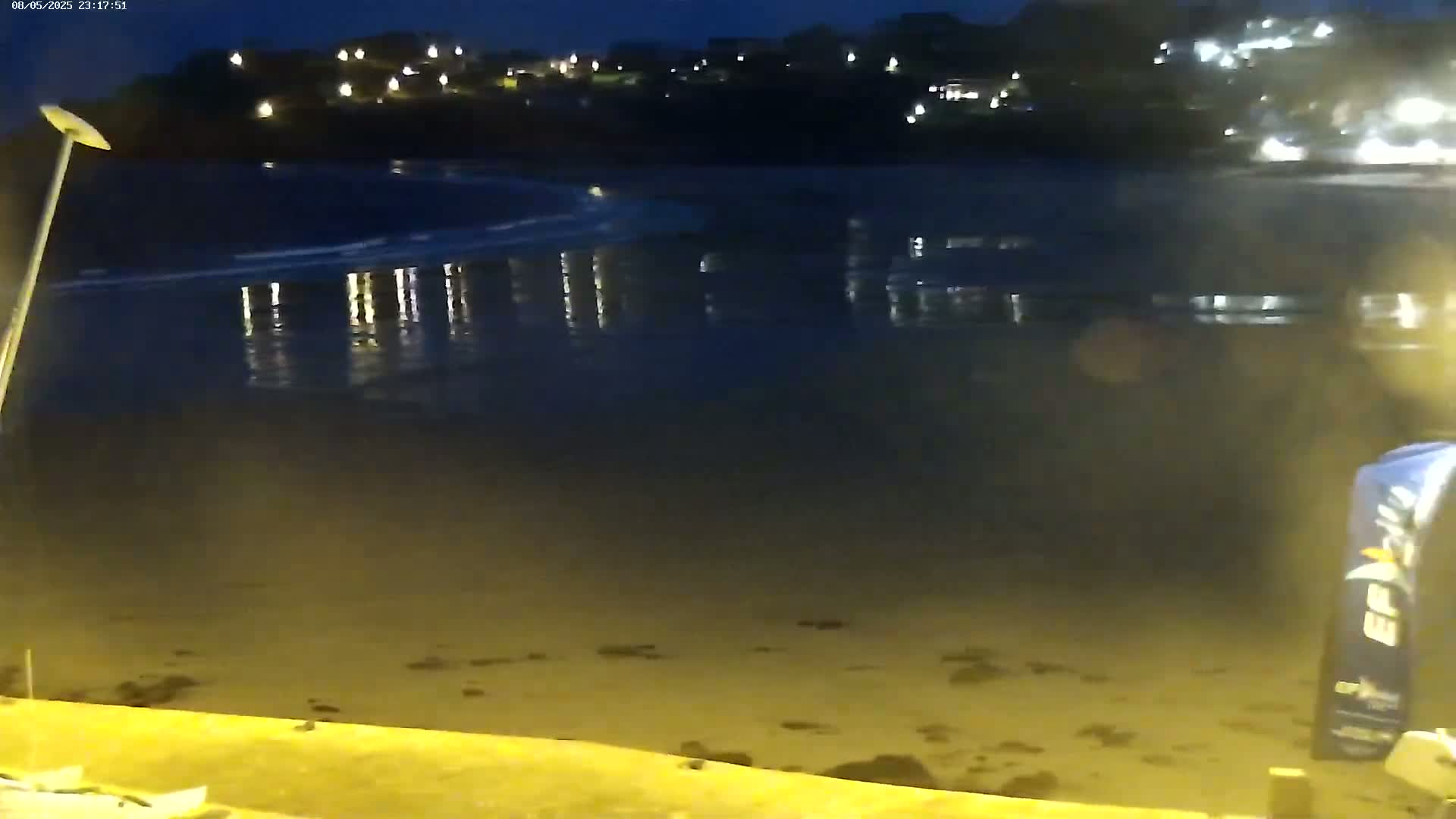 A dark, sandy beach at night is gently lapped by calm water reflecting the lights of a town in the distance.