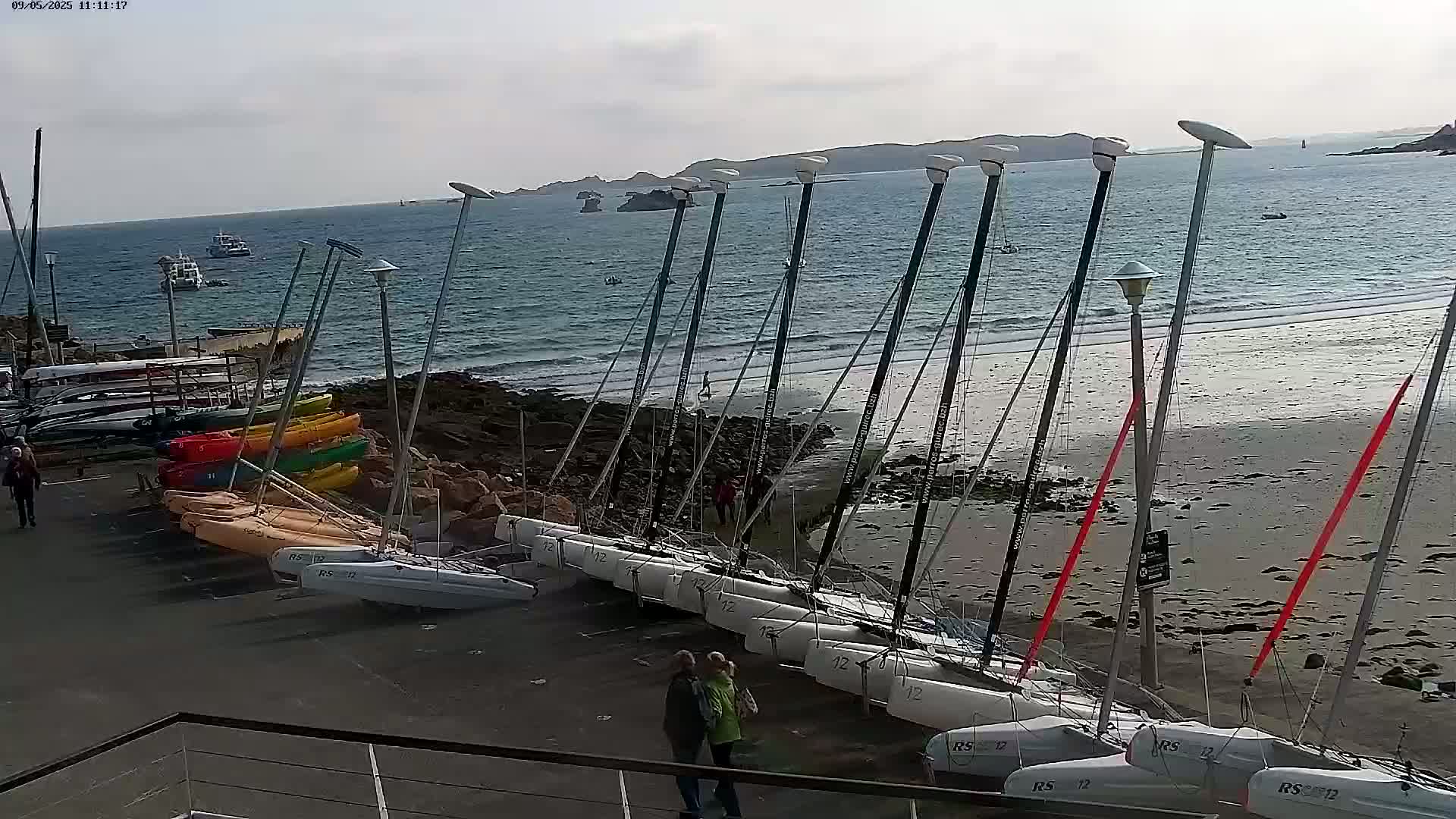 Several catamarans are beached on a sandy shore next to a rocky outcrop, with a calm sea and distant land visible under a mostly sunny sky.