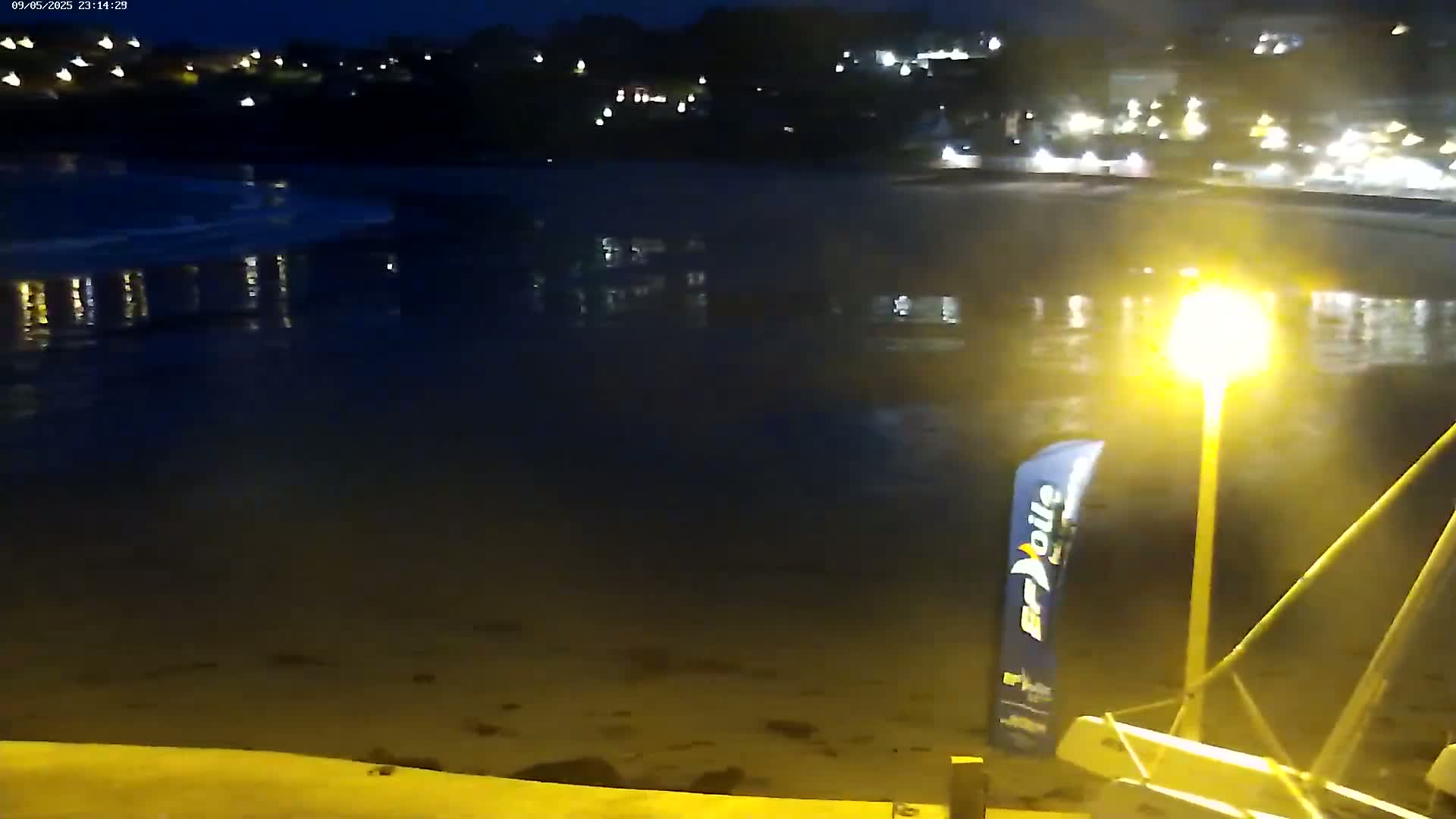 A nighttime view of a calm, dark body of water reflecting the lights of a town across a sandy beach, with a sailboat partially visible in the foreground under a bright lamppost.