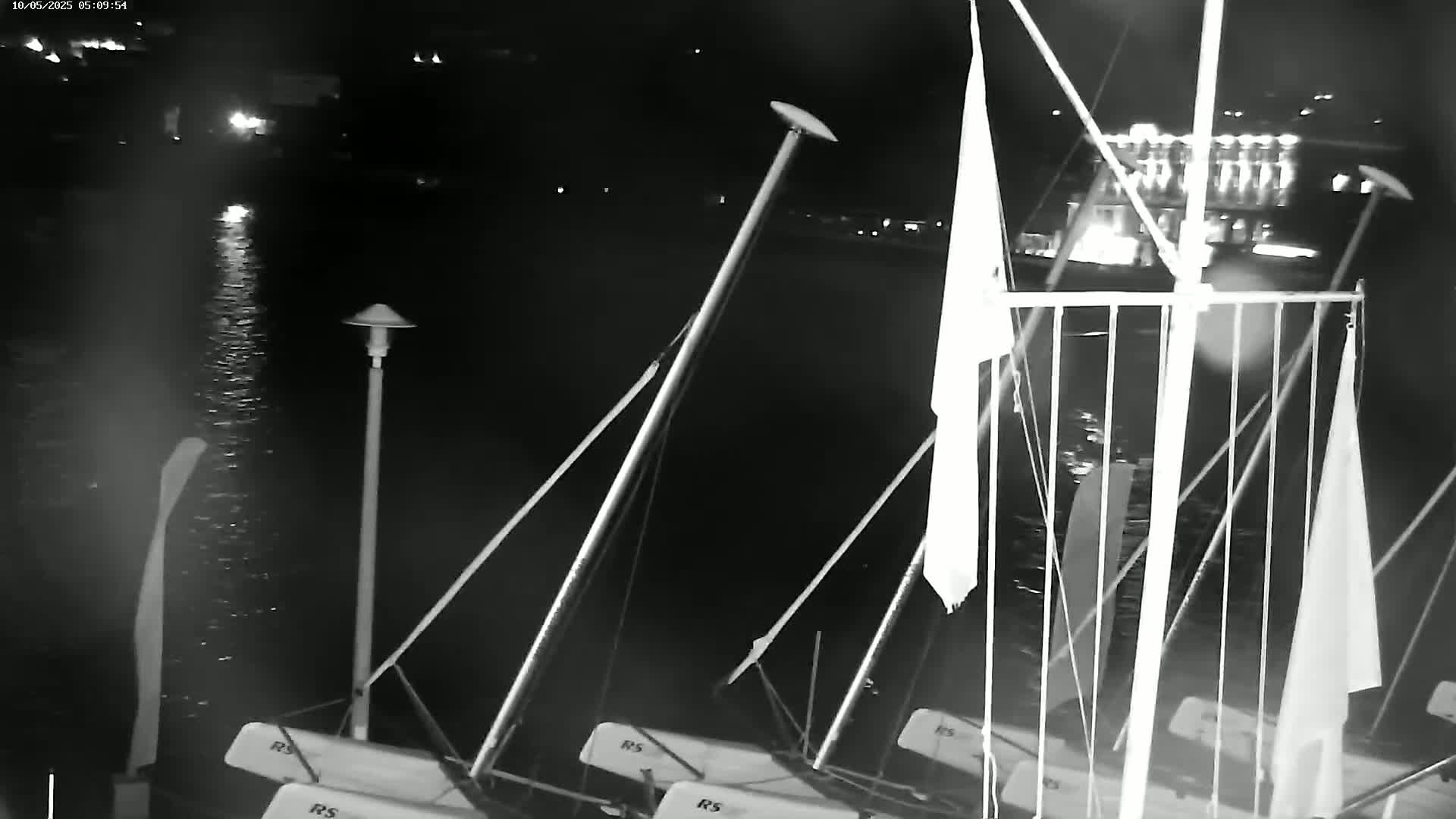 Several sailboats are moored at a dock at night, with calm water visible in the foreground and lights illuminating a building in the background.