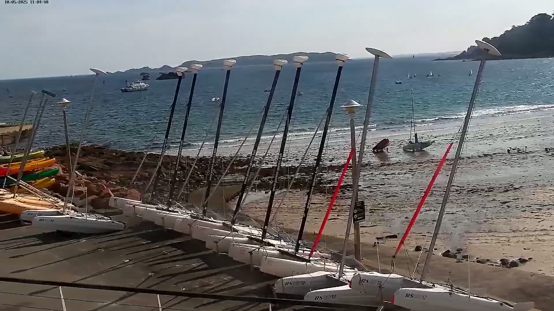 Many small sailboats are beached on a sandy shore near a rocky coastline under a sunny sky, with a few sailboats visible on the calm water.