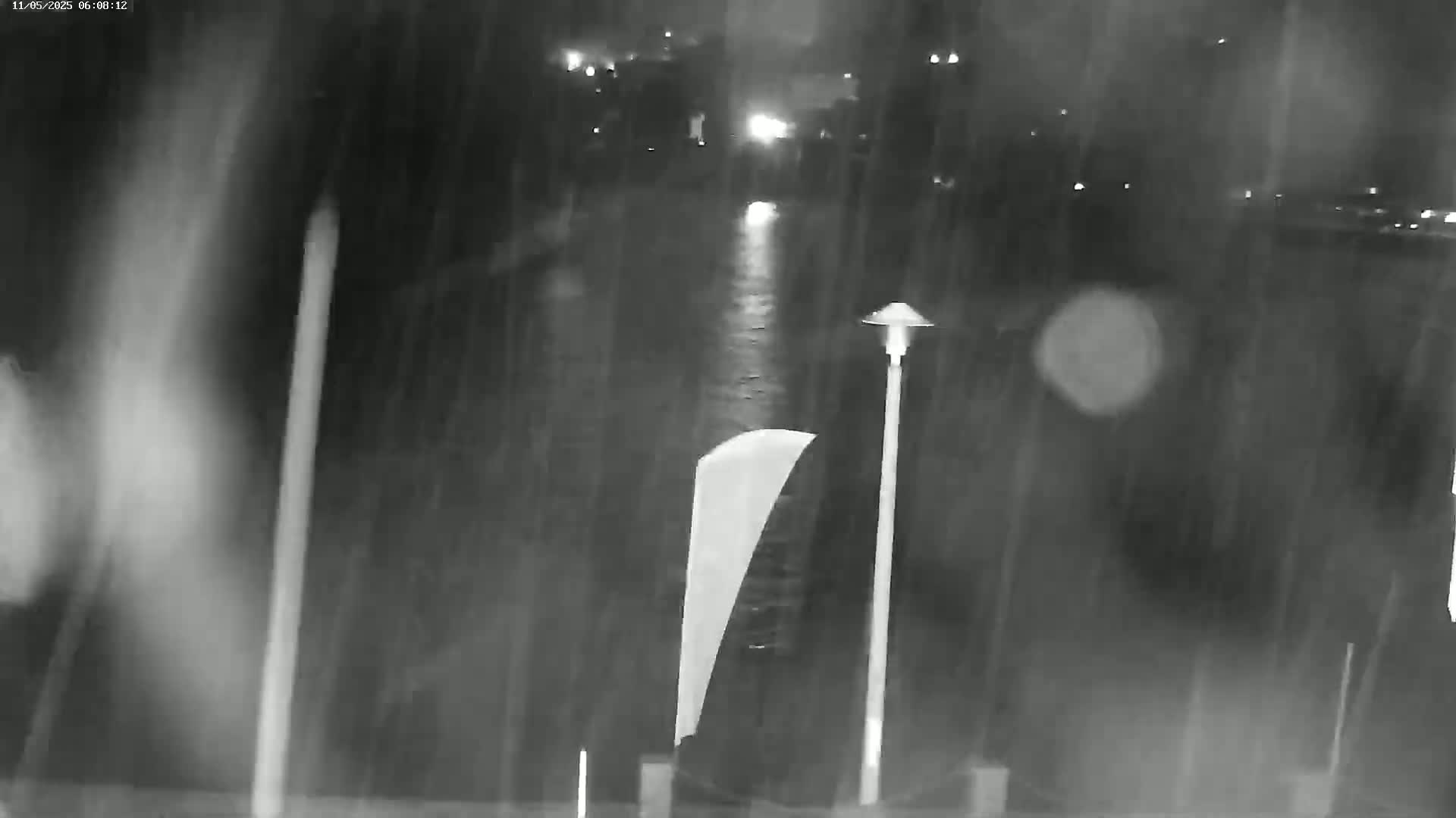 A nighttime, rainy scene shows a body of water reflecting lights from the opposite shore, with a tall lamppost and a partially visible, light-colored object in the foreground.