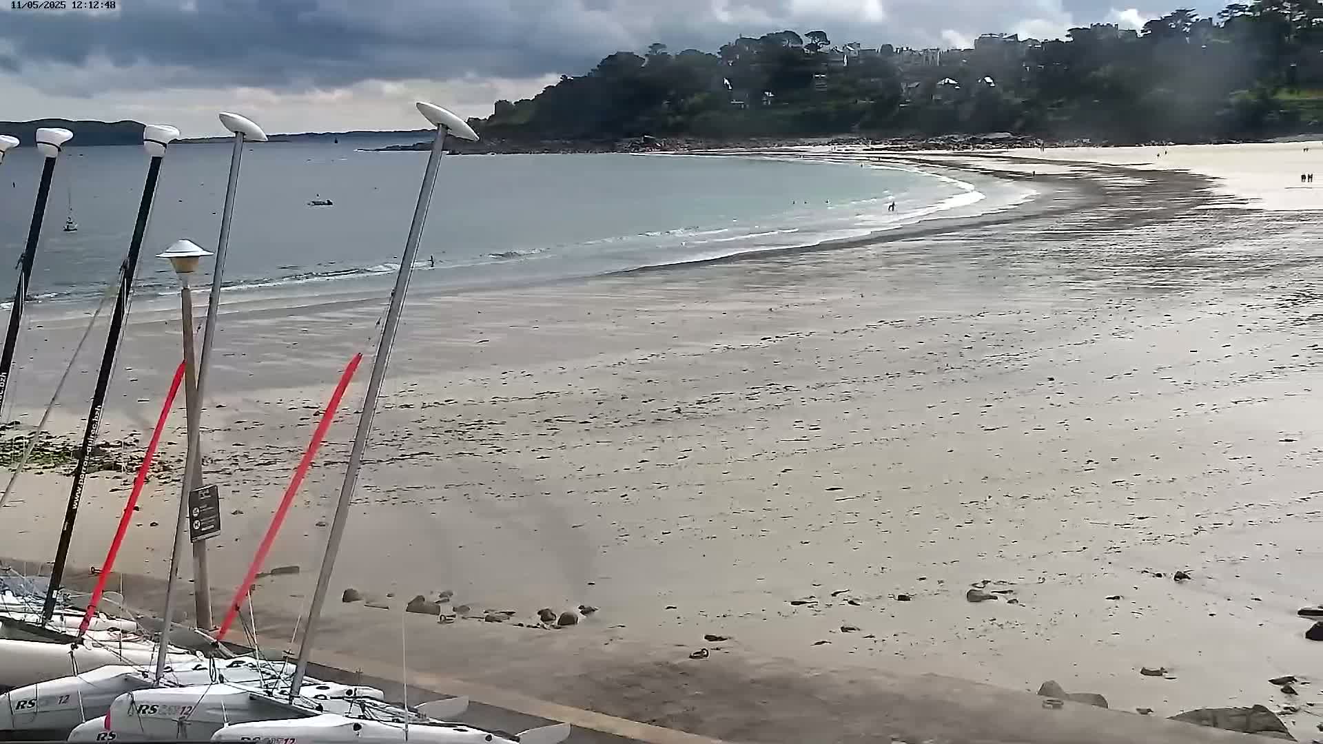 Several sailboats are docked on a sandy beach under a cloudy sky, with a calm sea extending to a distant shoreline.