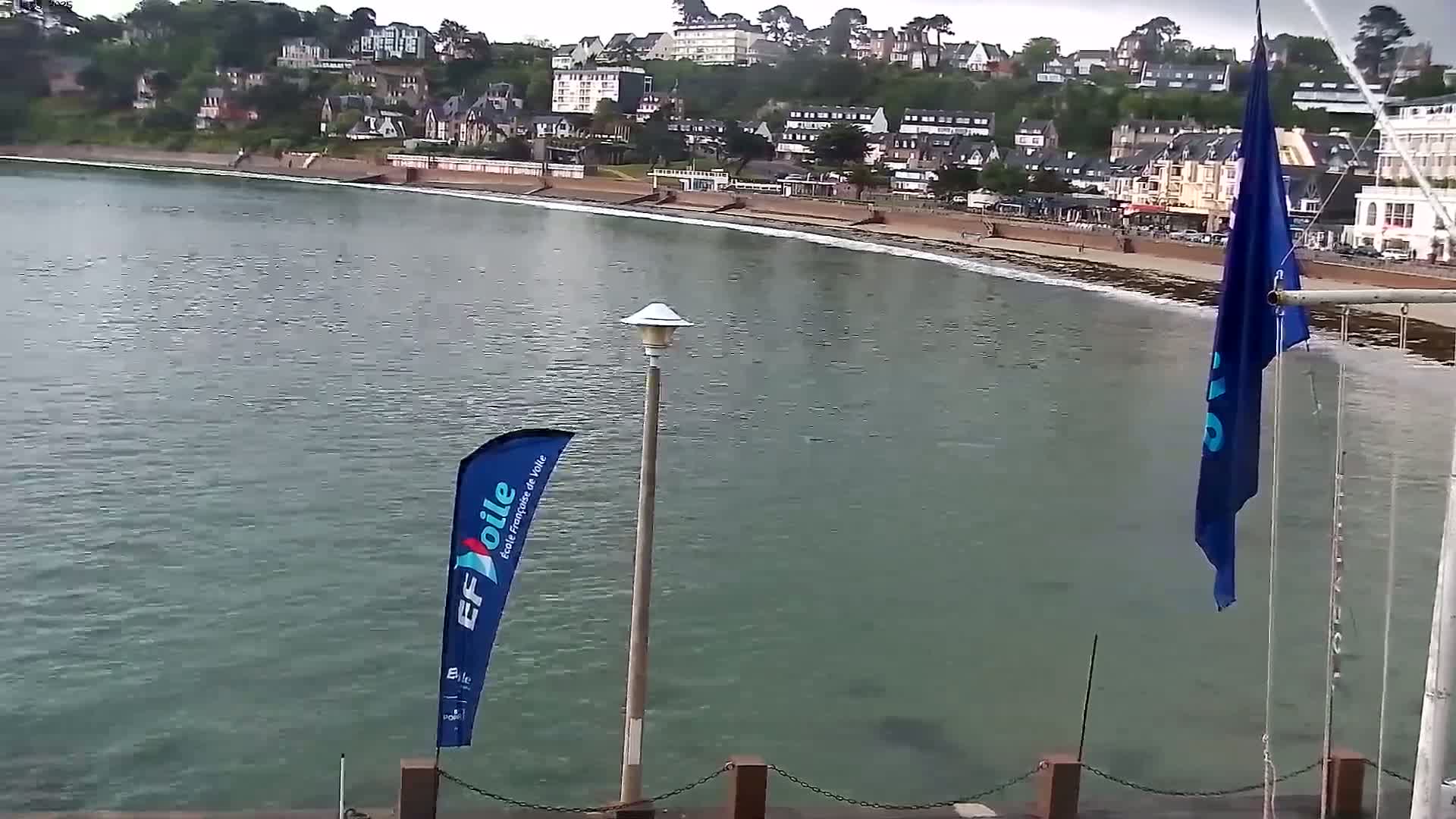 A calm body of water laps a sandy beach in front of a hillside town under an overcast sky.