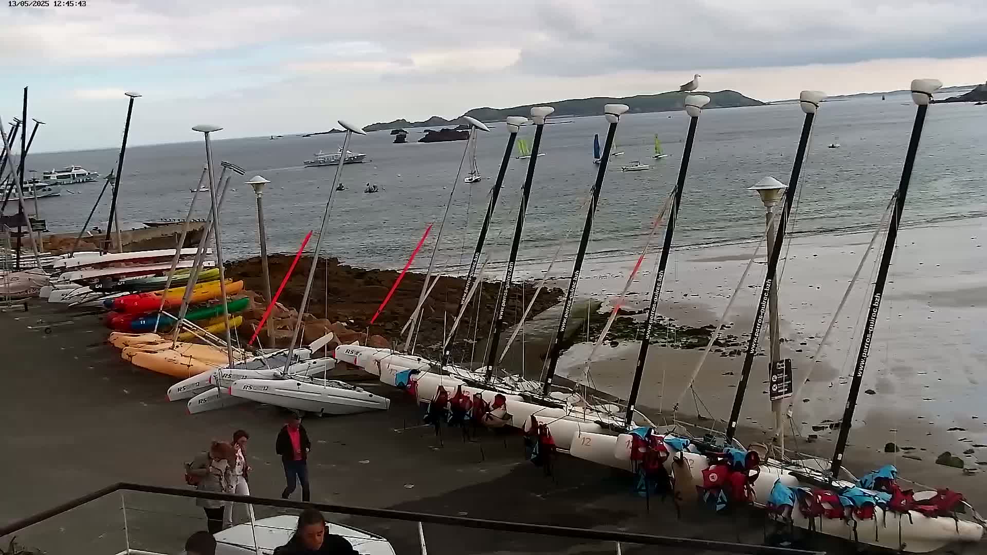 Numerous sailboats and kayaks are beached on a sandy shore near rocky outcrops, under an overcast sky.