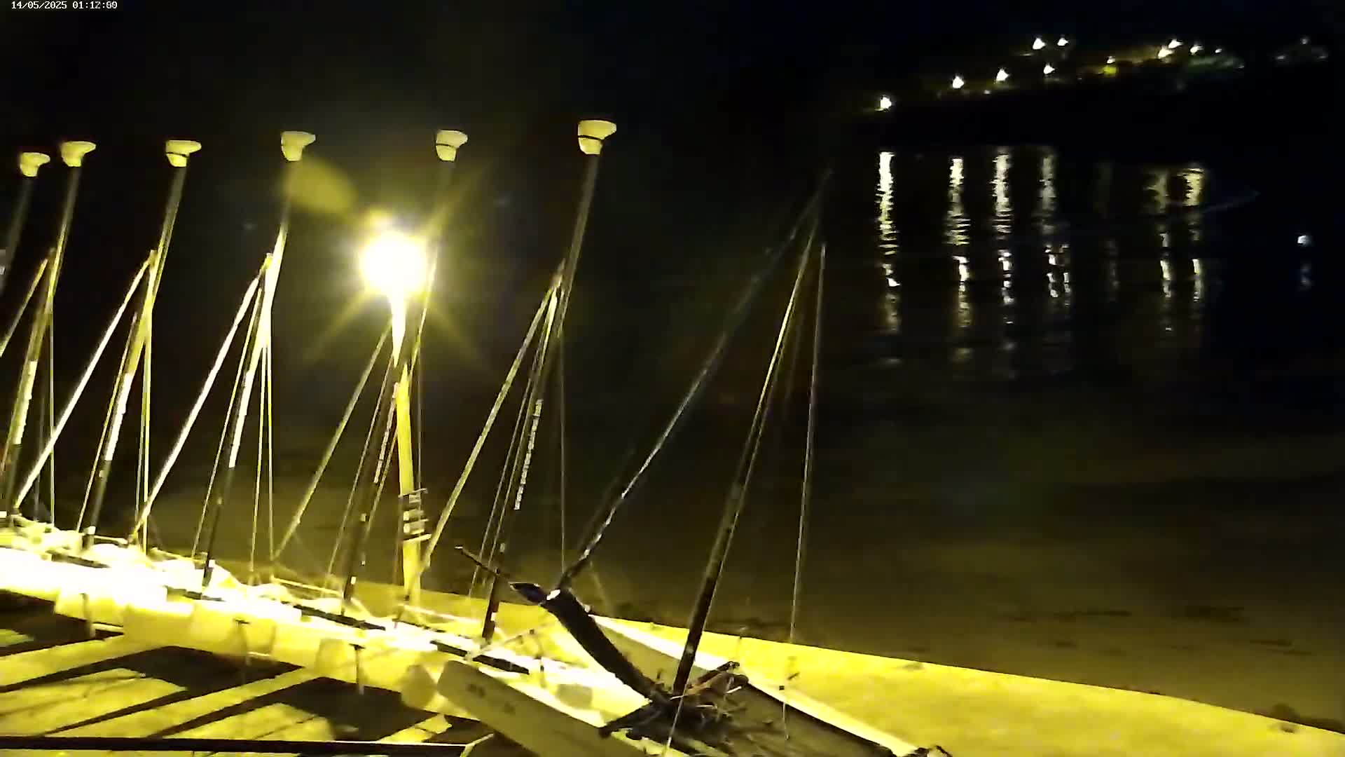 At night, several small sailboats are beached on a sand-colored shore illuminated by a nearby lamppost, with calm water reflecting lights in the background.