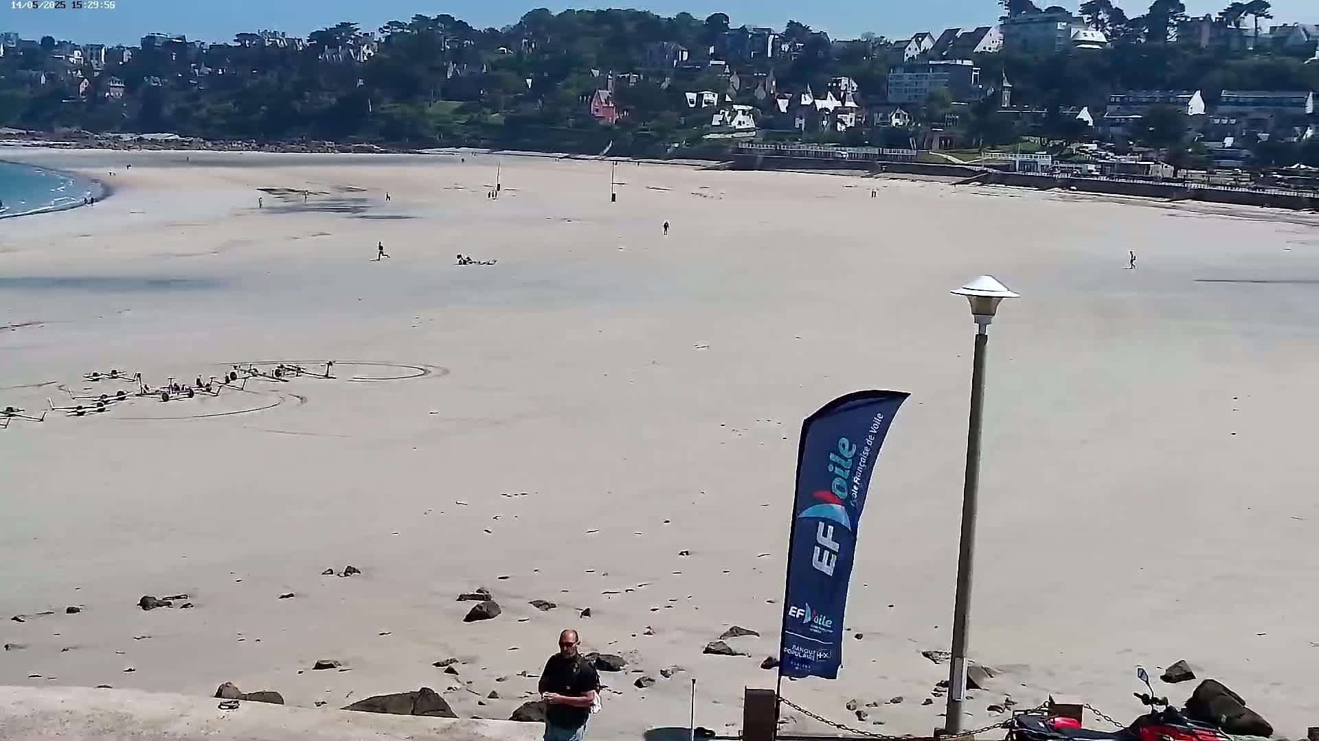 A mostly empty sandy beach stretches below a line of buildings on a sunny day, with a few people scattered about and some equipment arranged in circles on the sand.