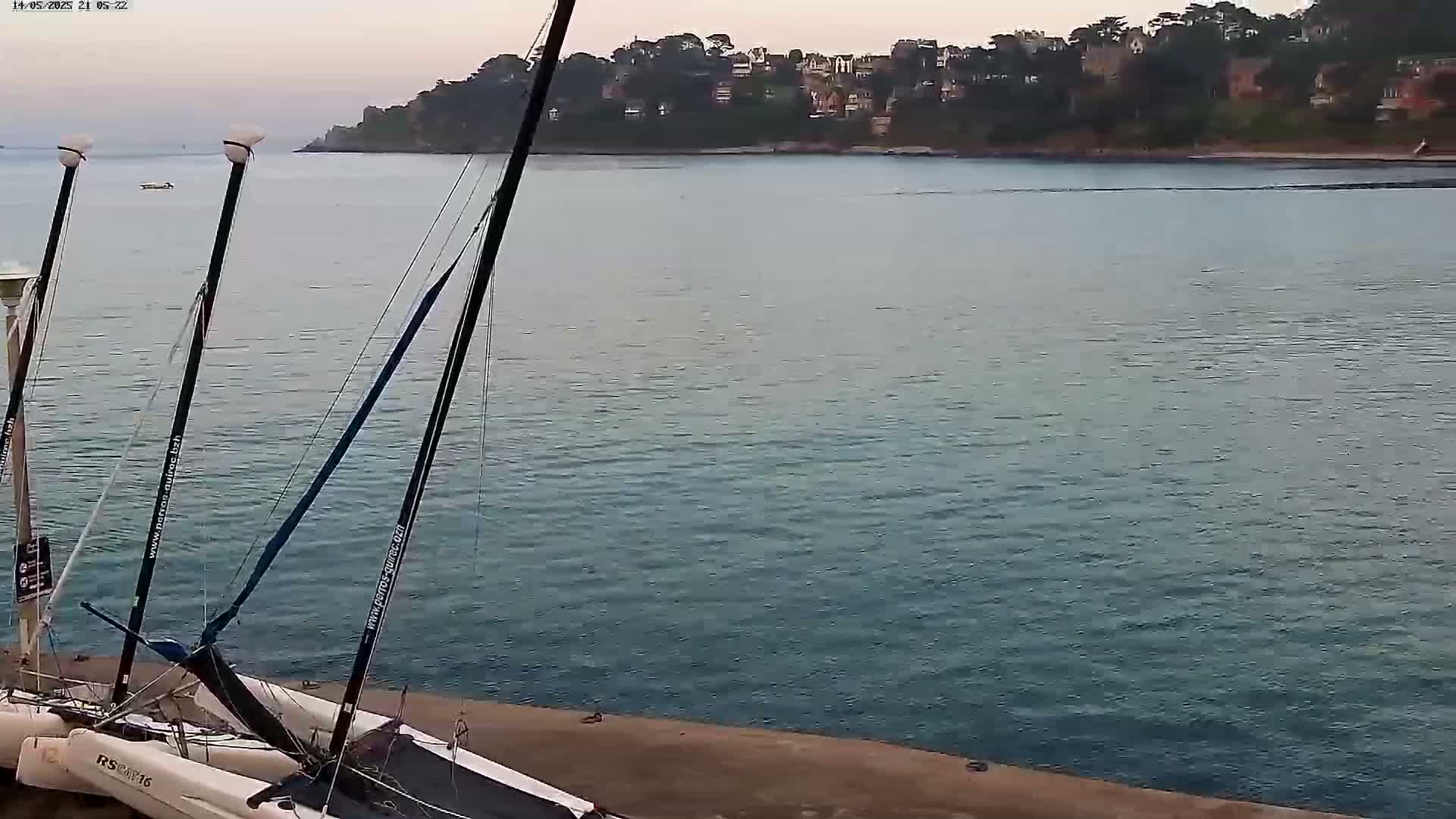 Two catamarans are docked at a waterfront, with calm water and a distant shoreline featuring houses and trees under a dusky sky.
