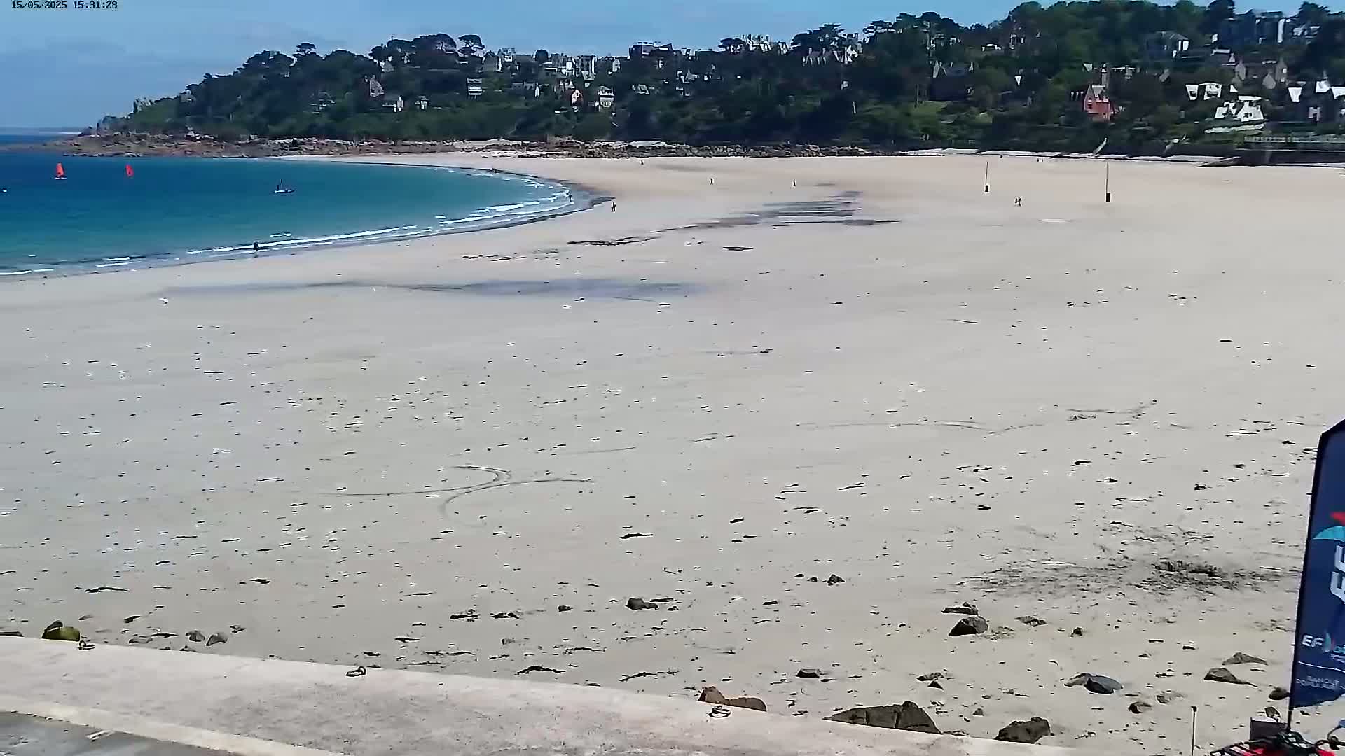 A wide, sandy beach stretches towards a calm, light-blue ocean under a clear, sunny sky, with a few sailboats visible in the distance and a small town nestled on the hill overlooking the beach.