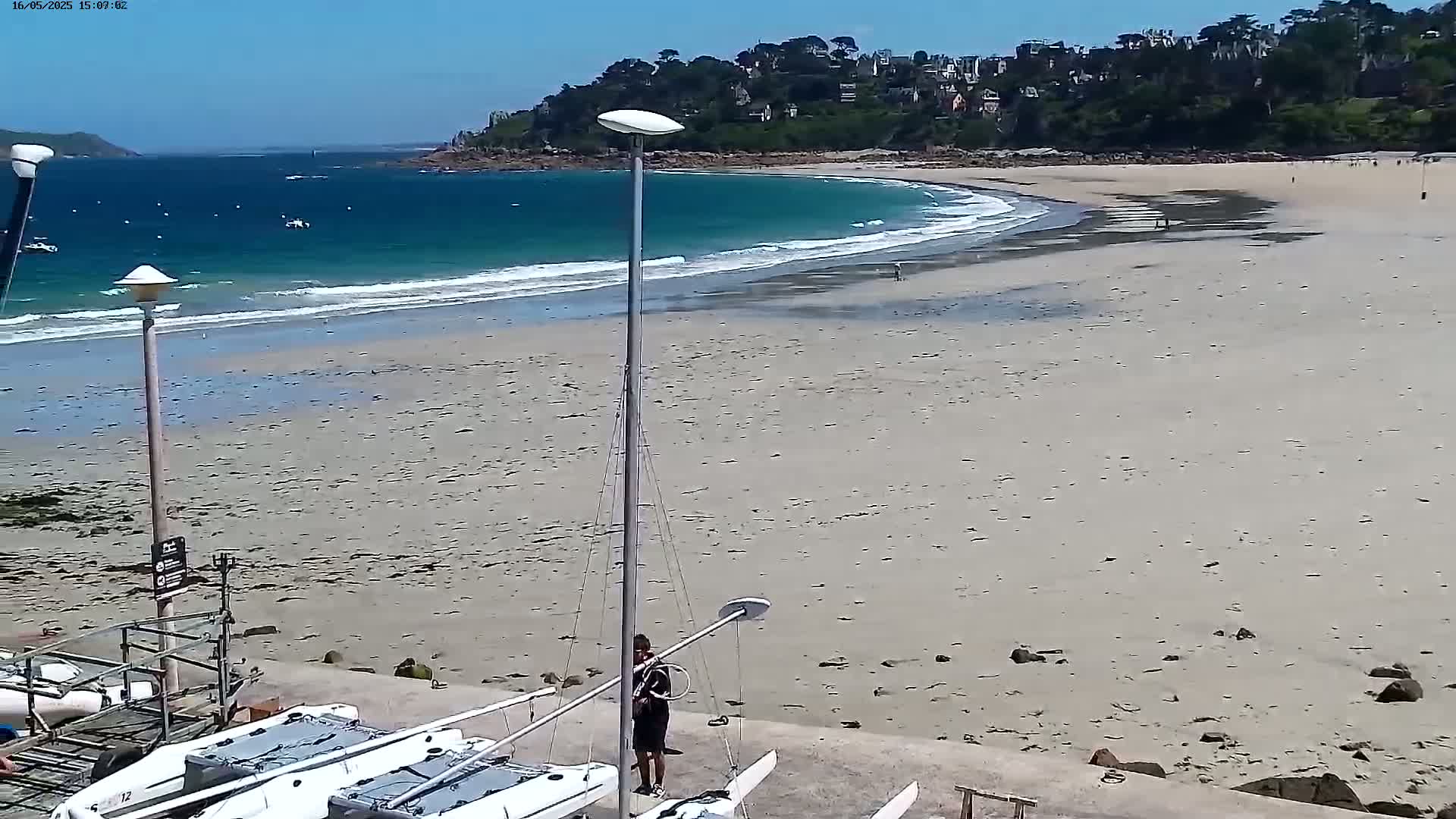 A sandy beach with calm waves laps at the shore under a sunny sky; a person stands near several small sailboats on the beach near a rocky outcropping and a village in the distance.