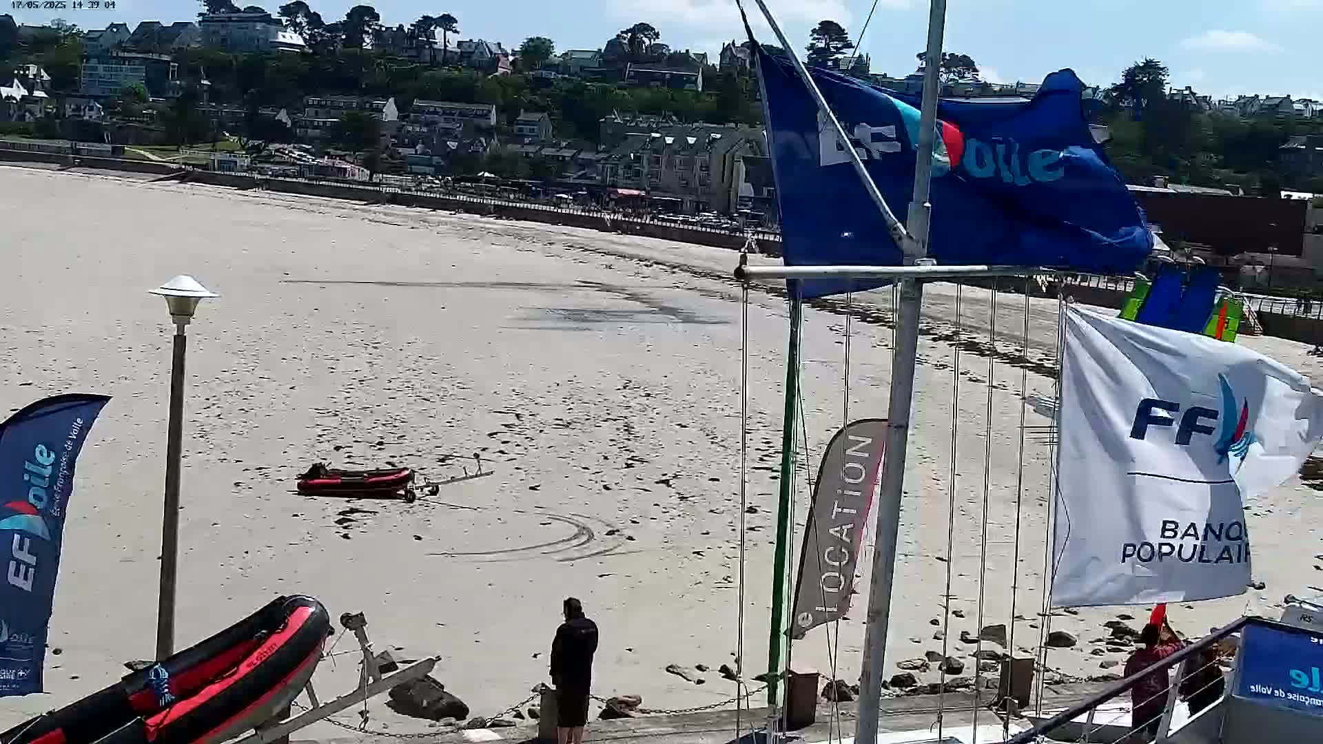 A sandy beach with two small inflatable boats, a person standing near a boat trailer, and a collection of flags on poles are visible on a sunny day, with a town in the background.