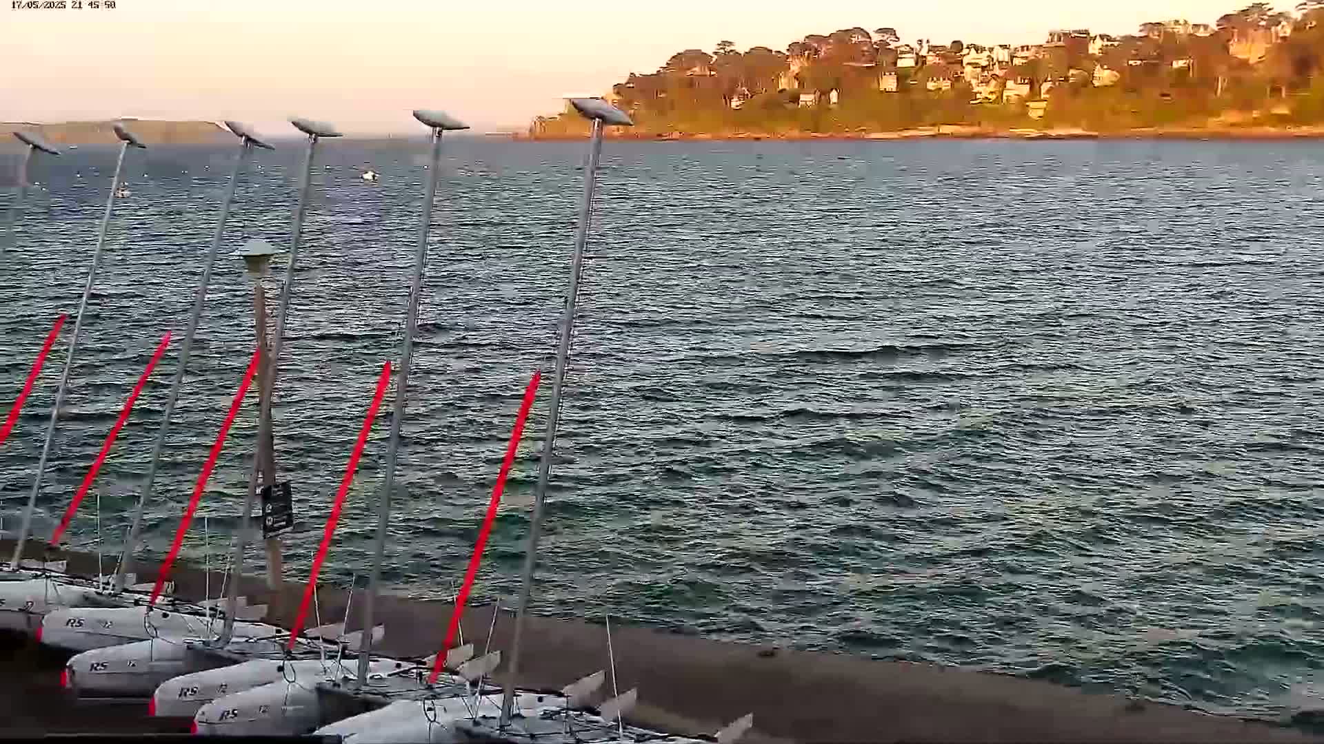 Several small sailboats are docked at a shoreline next to a body of water with small waves under a partly cloudy, twilight sky.