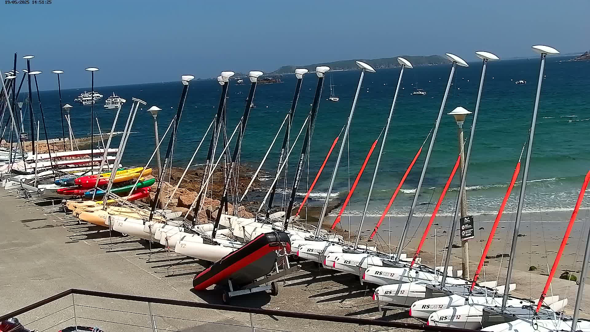 Numerous small sailboats and kayaks are lined up on a beach next to a calm, blue ocean under a clear, sunny sky.