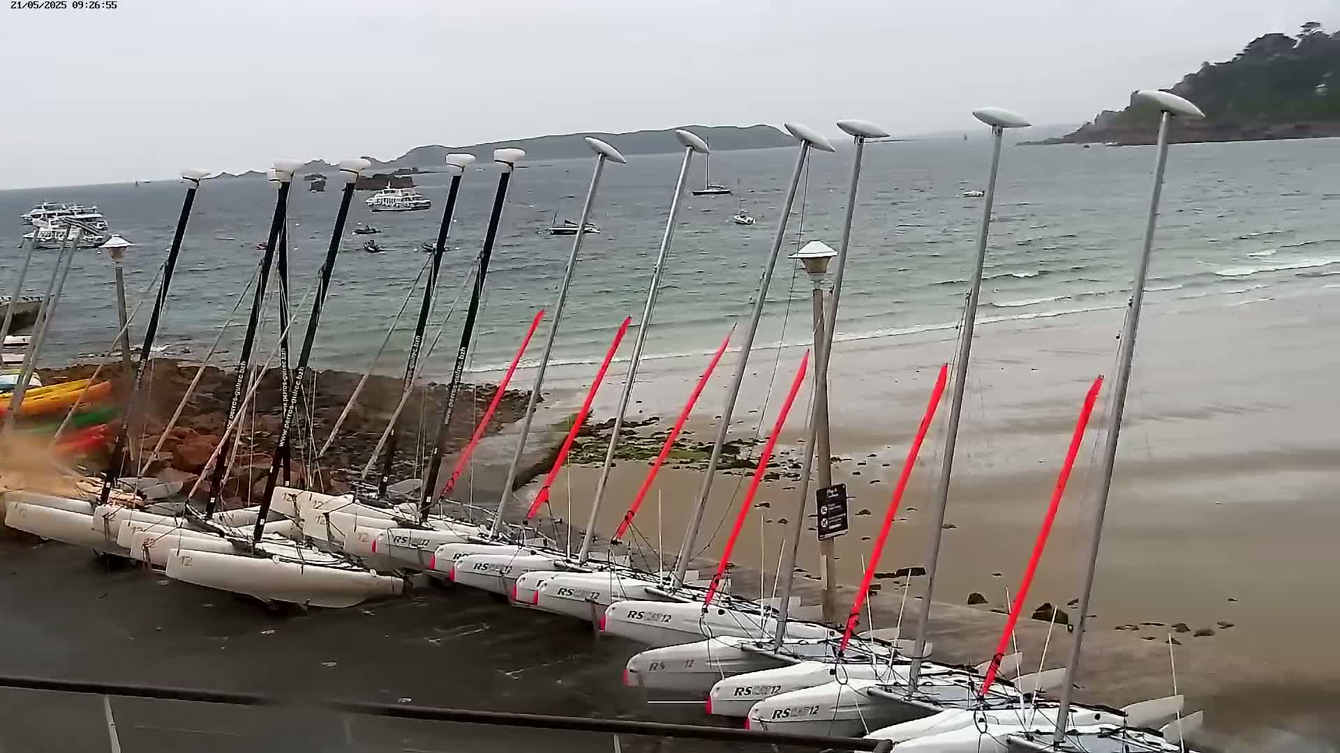 Several catamarans are beached on a sandy shore next to a rocky outcrop, with choppy water and overcast skies.