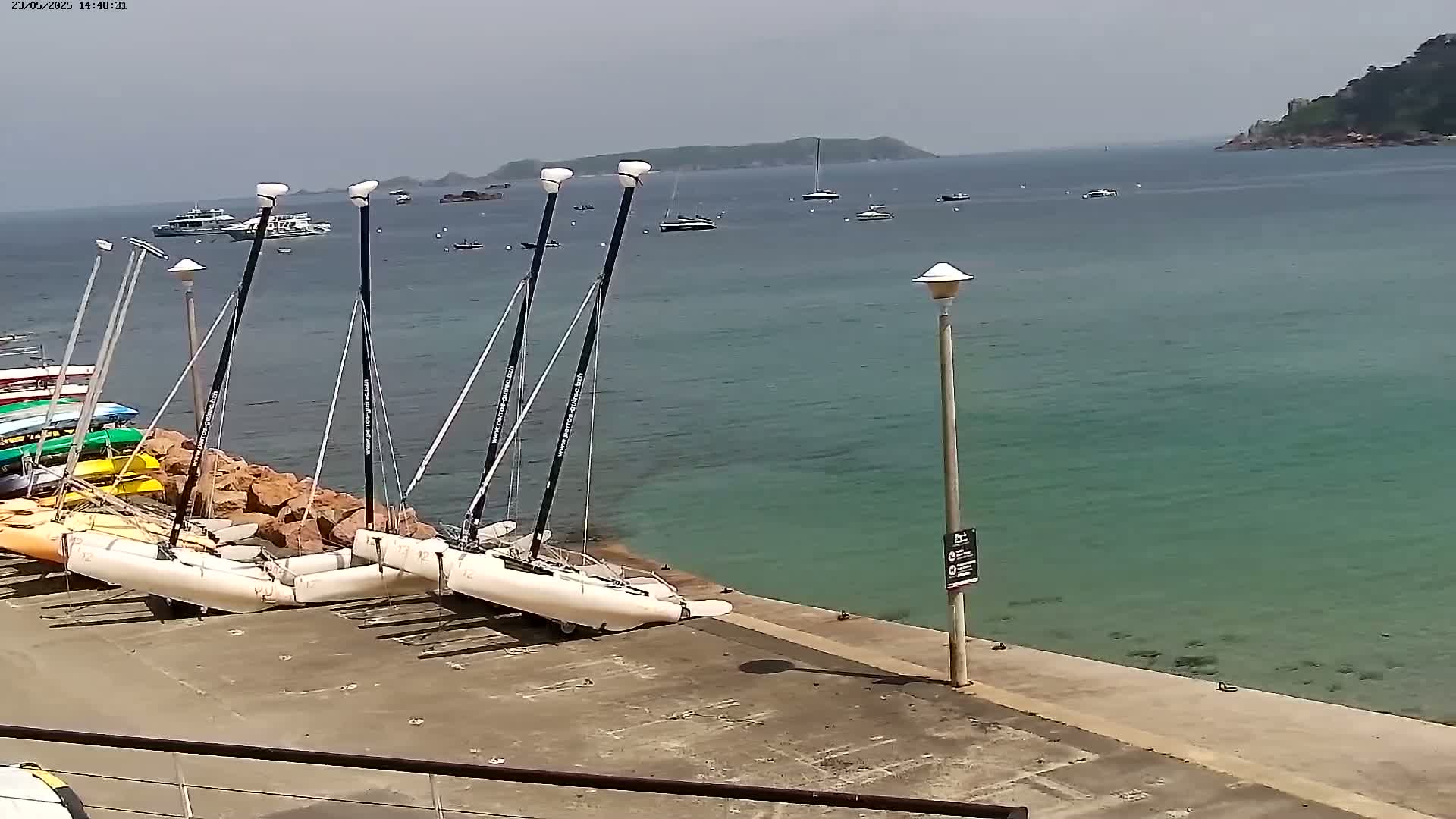 Several catamarans are docked near a rocky shore next to calm, teal-colored water under a mostly clear sky.