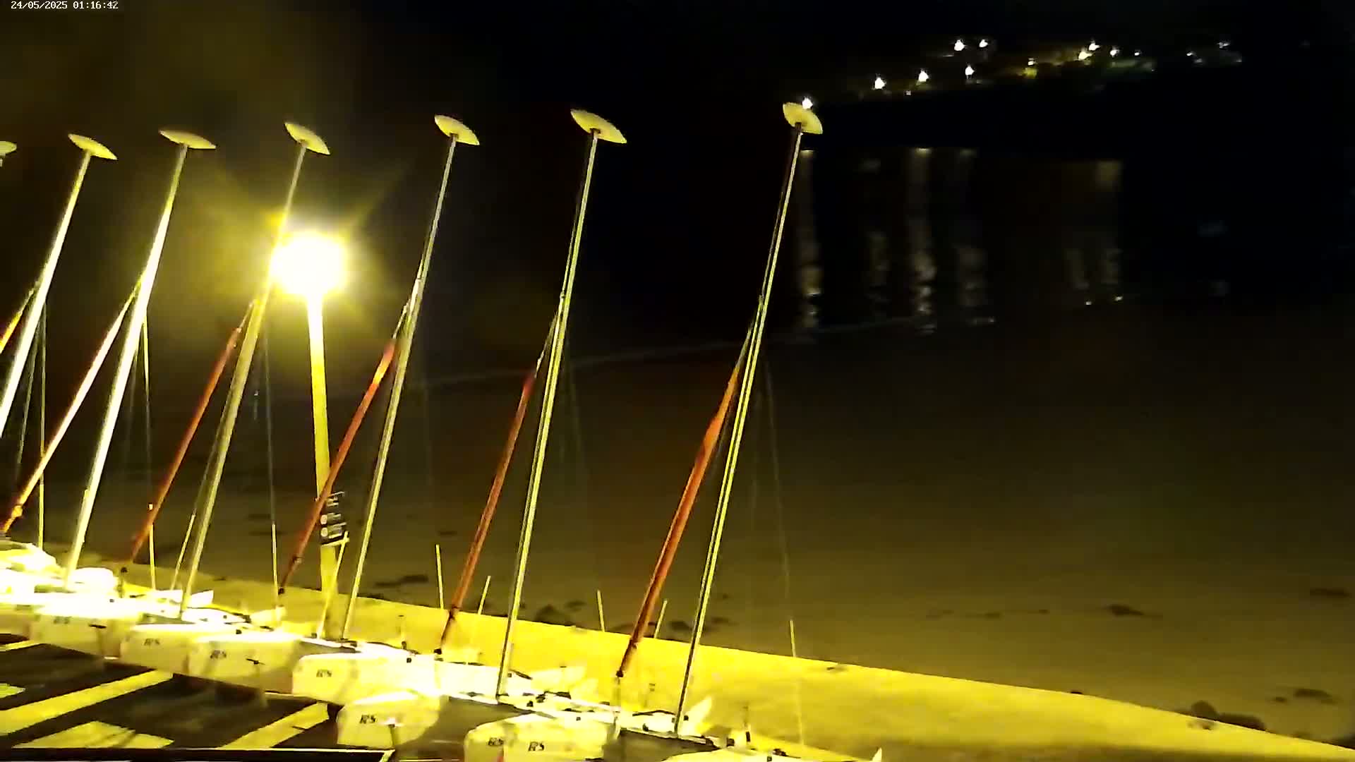 Several sailboats are docked at a shore at night under a dark sky illuminated by artificial light.