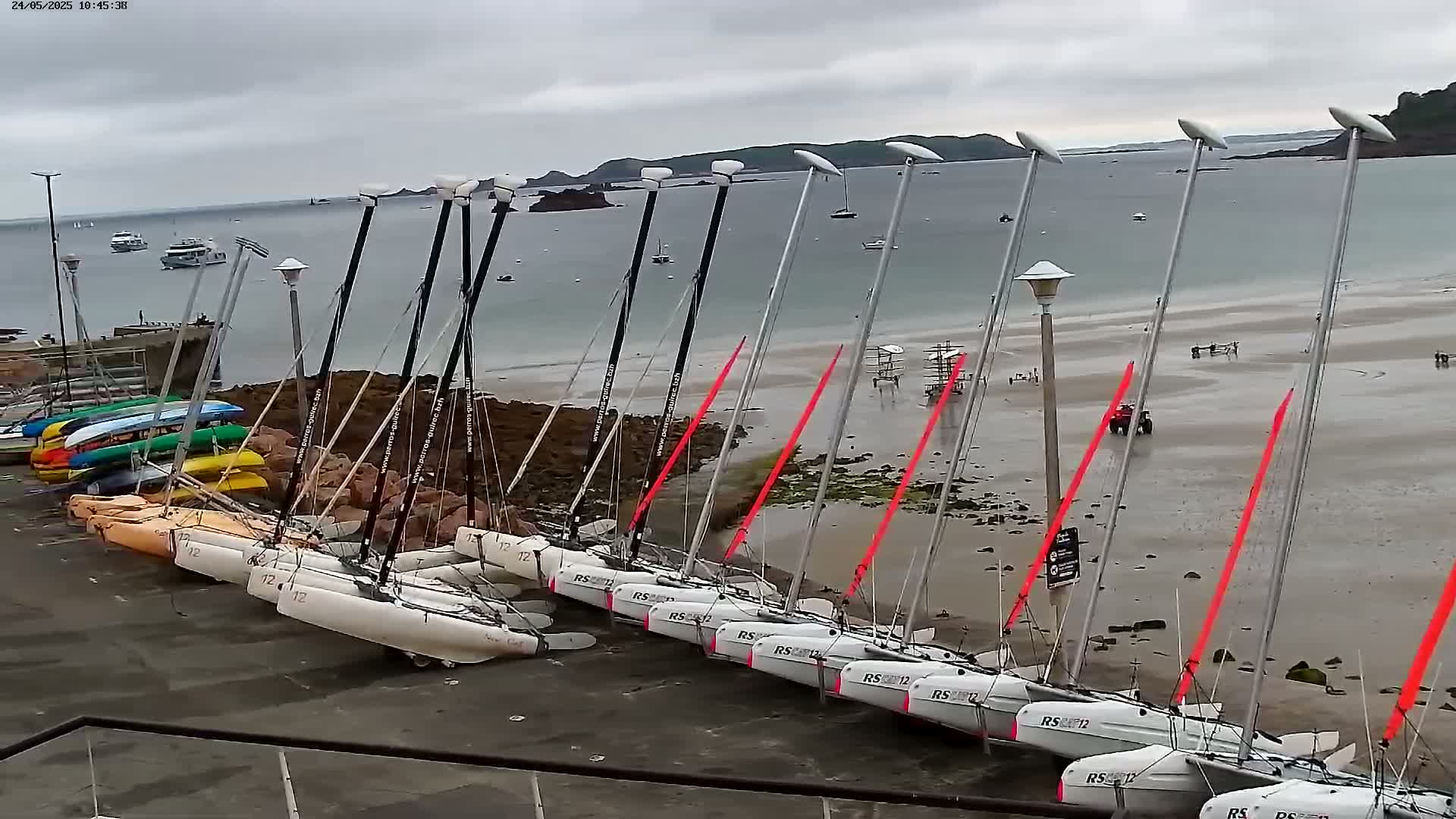 Several catamarans are beached on a sandy shore under an overcast sky, with other boats visible in the calm water beyond.