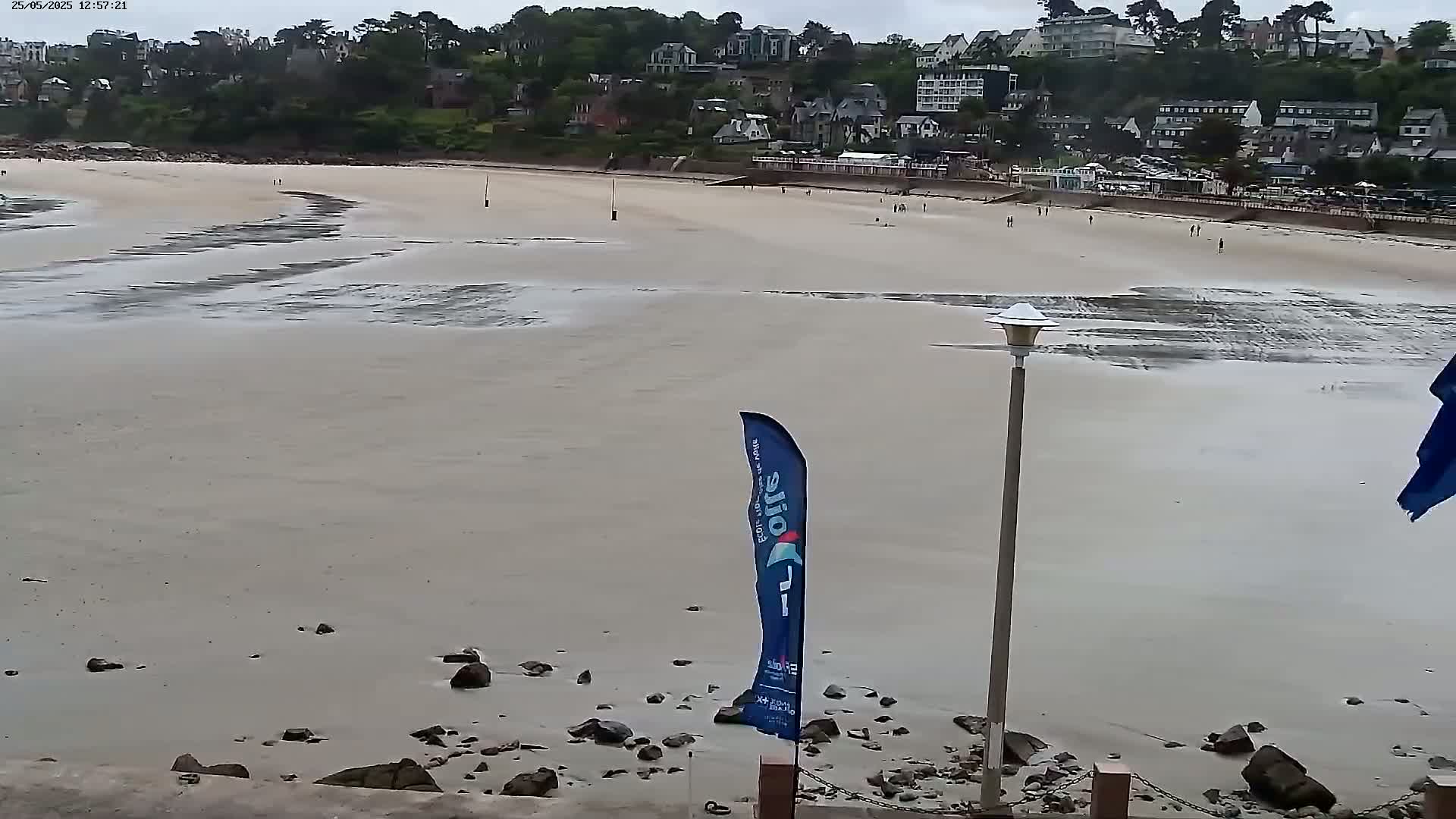 A sandy beach with low tide, revealing exposed sand and scattered rocks, stretches out before a hillside town under an overcast sky.