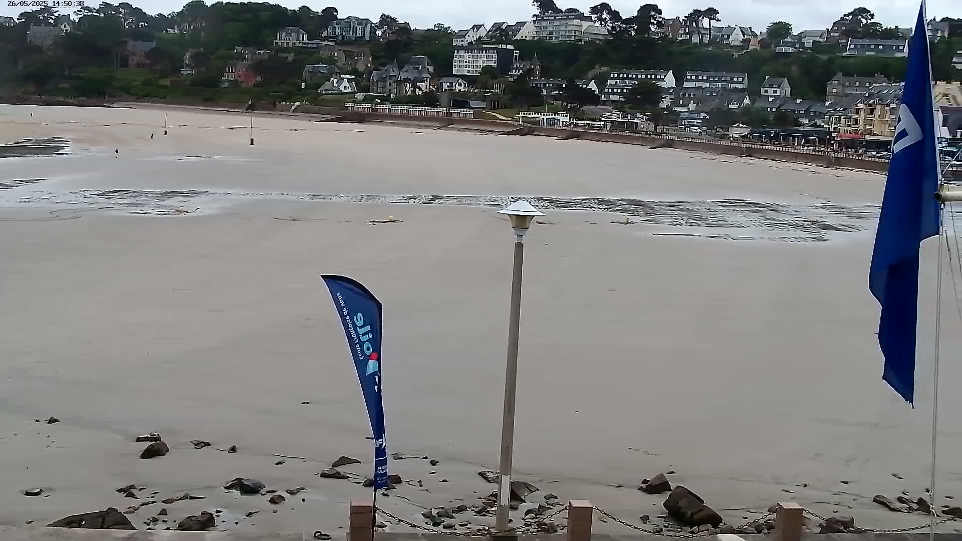 A mostly exposed sandy beach with a few rocks and a lamppost, overlooked by a row of buildings under a cloudy sky.