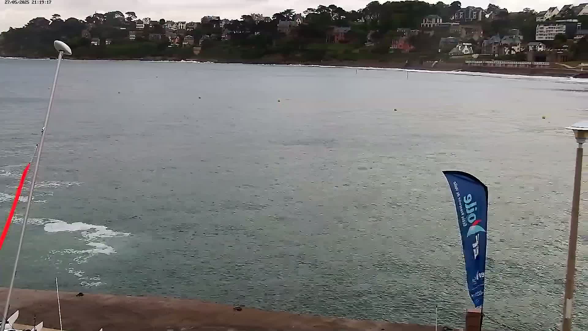 A body of water with a sailboat and a flag visible near a shoreline with houses on a hill in the background under an overcast sky.