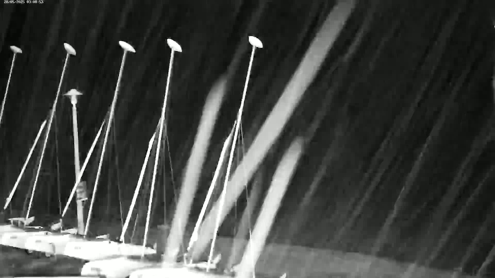 Several sailboats are moored in a harbor during a heavy downpour at night.