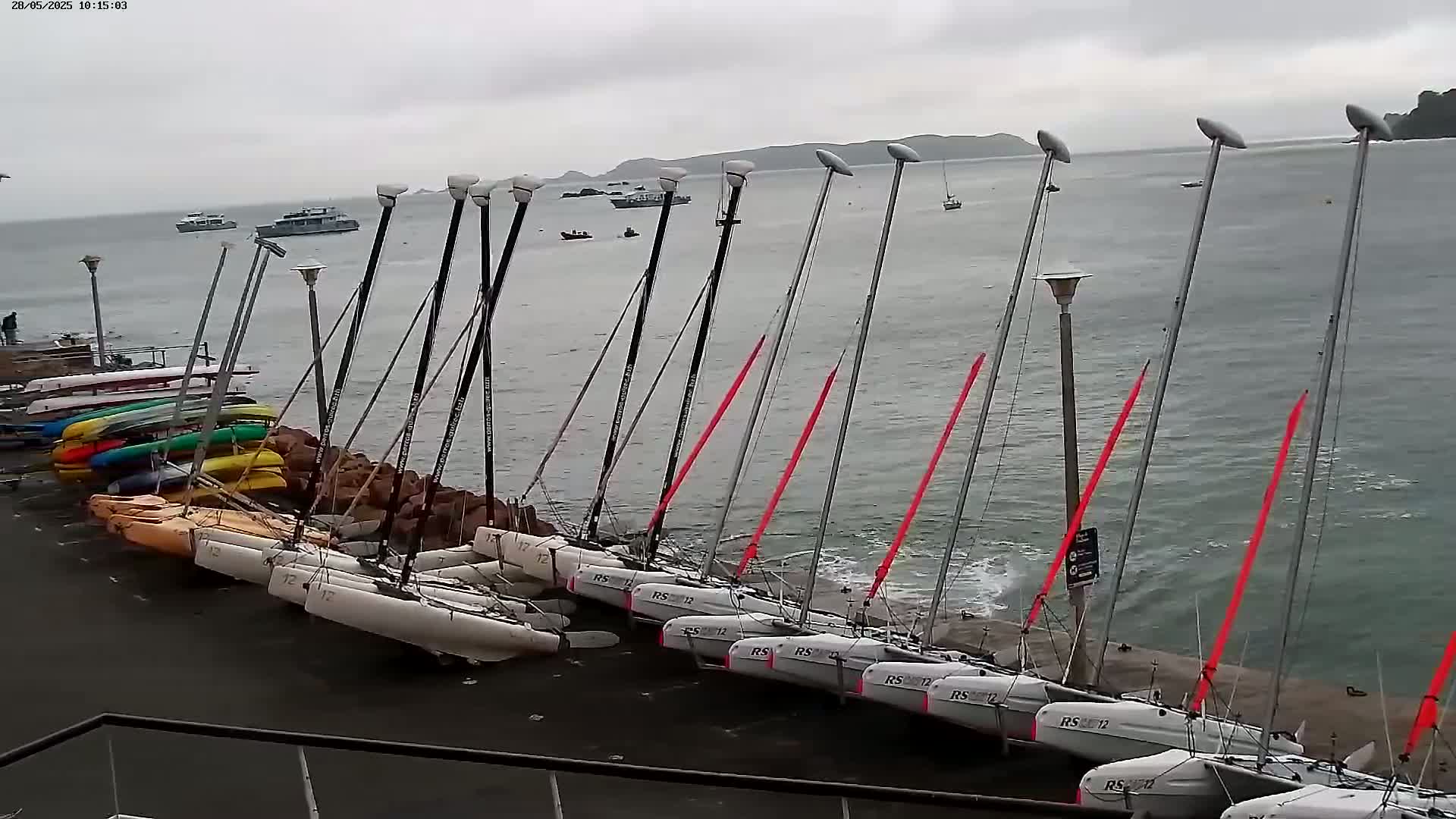 Several catamarans and kayaks are moored along a rocky shoreline on an overcast day, with a few boats visible in the distance on choppy water.