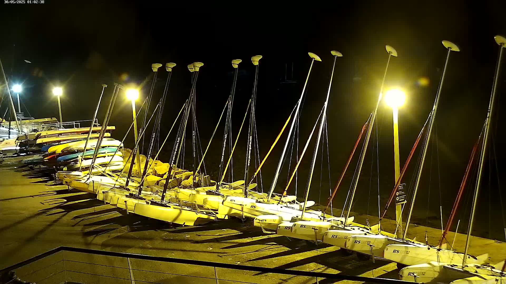 Many sailboats are docked at night under bright artificial lights.