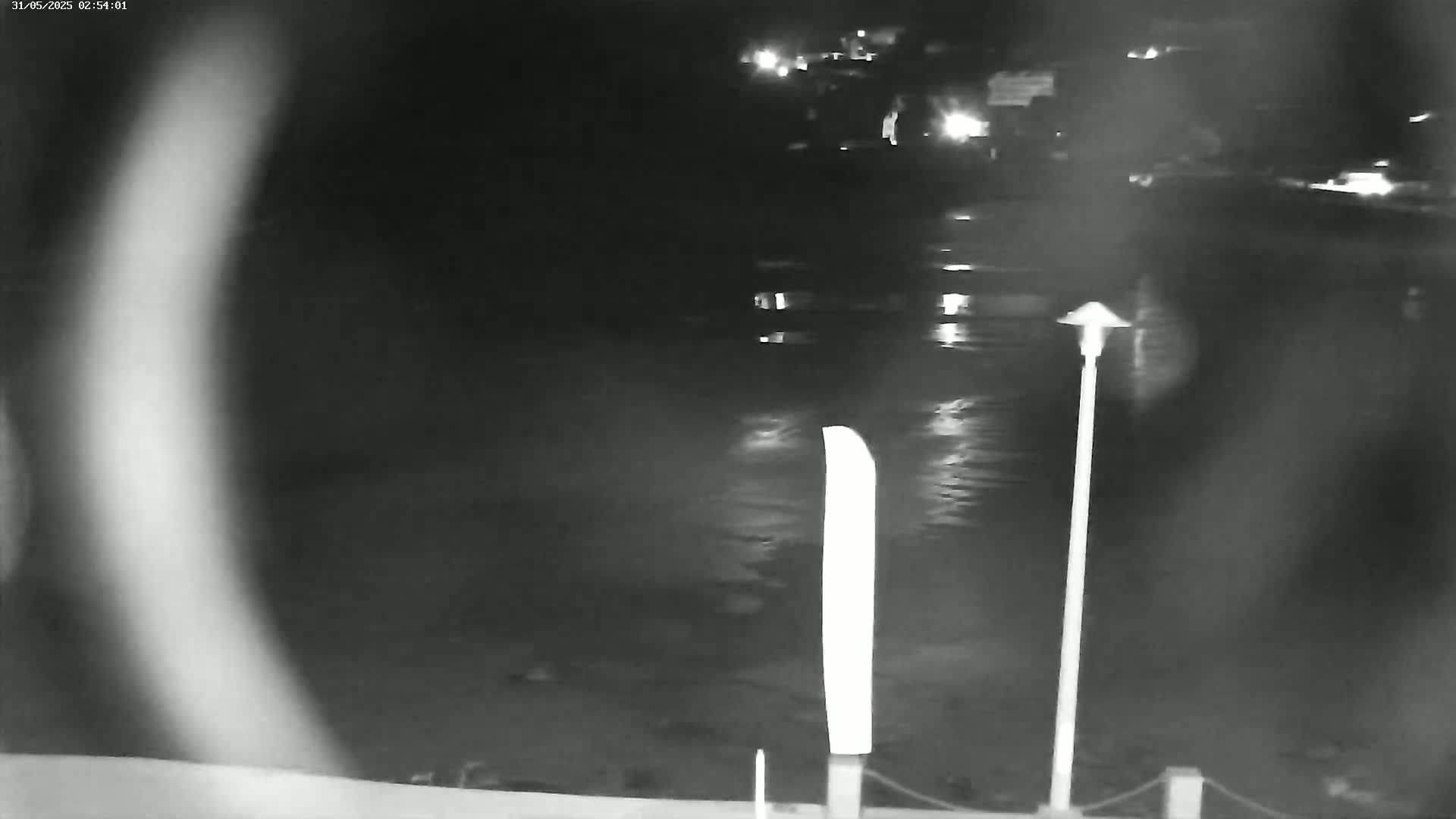 A nighttime, low-light image shows a dark body of water reflecting lights from the distance, with a tall, thin, light-colored post in the foreground.