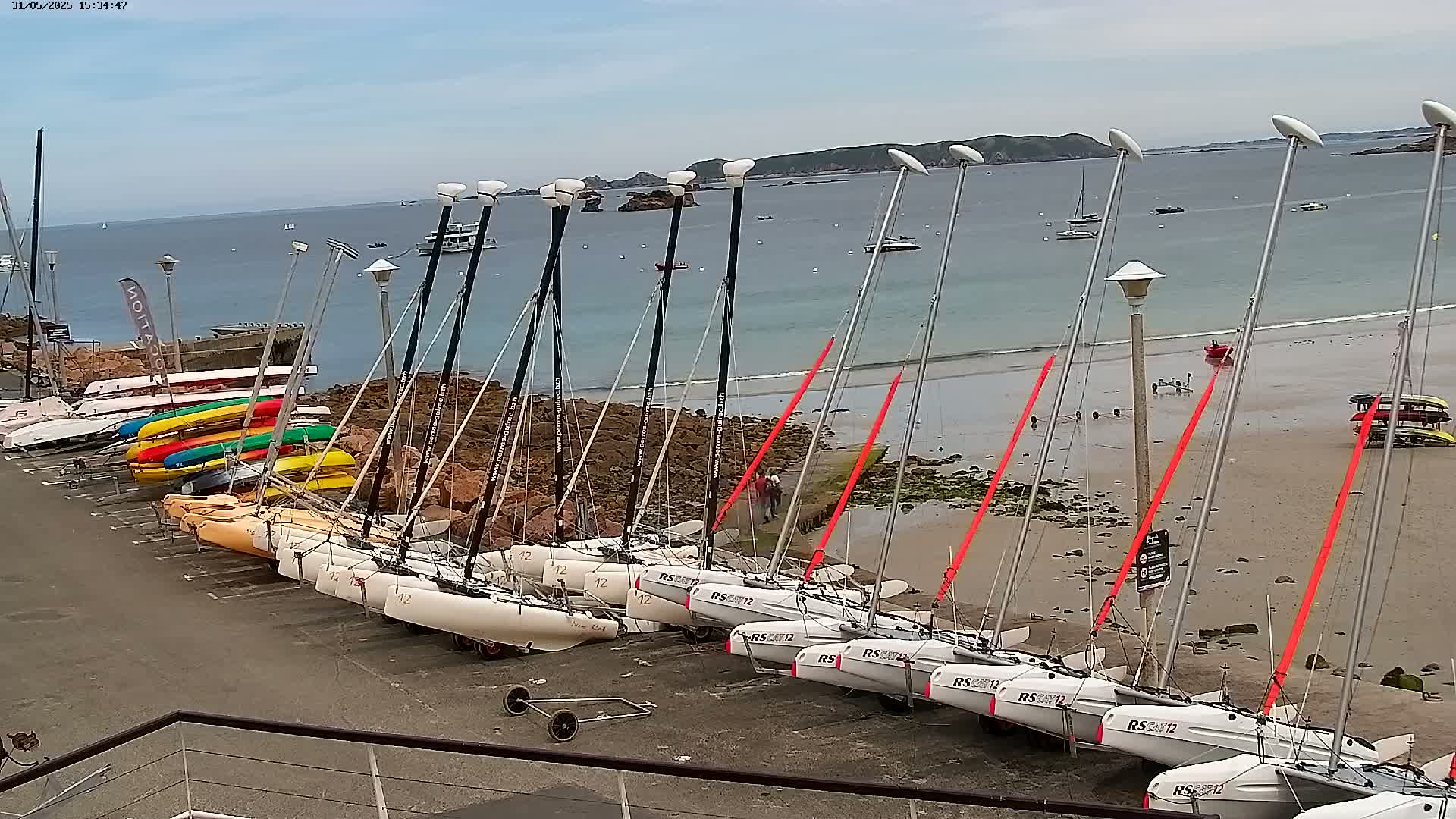 Numerous sailboats and kayaks are parked on a beach near a calm body of water under a partly cloudy sky.
