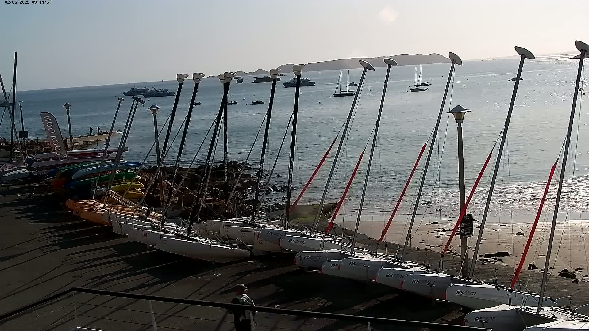 Many sailboats are beached on a sandy shore near calm, blue water under a clear sky.