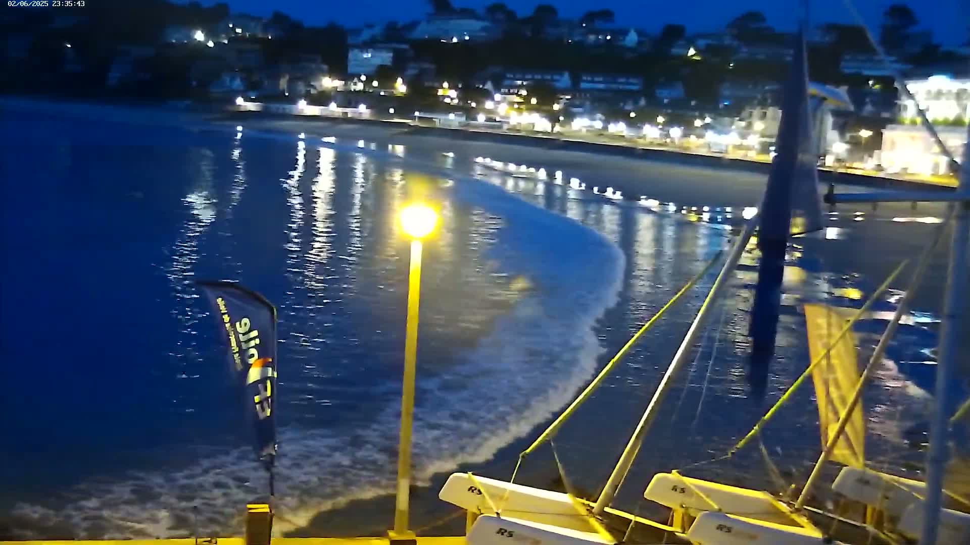Small sailboats are moored near a beach at night, with gentle waves lapping at the shore under a dark sky.