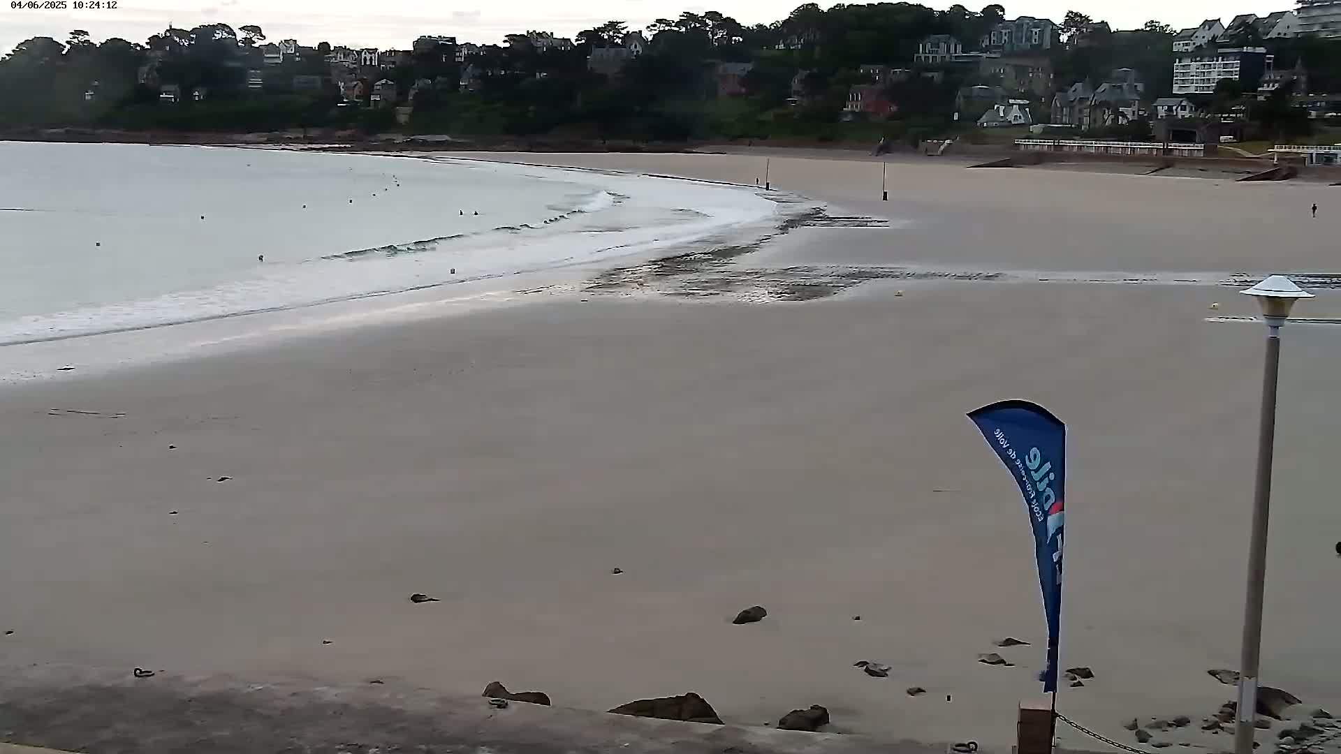 A sandy beach with gentle waves lapping the shore, under an overcast sky, is backed by a line of buildings and trees.